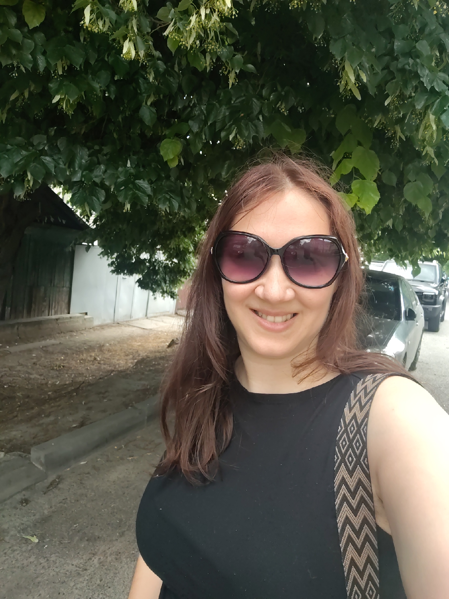 The image shows a woman taking a selfie outdoors. She has long, brown hair and is wearing large, dark sunglasses and a black sleeveless top with a decorative strap. The background features a tree with green leaves and small light-colored blossoms, suggesting it is spring or summer. To the left of the woman, there is a wooden fence and a light-colored wall, indicating a residential or rural setting. Behind her, two vehicles are visible, a car and a larger SUV or truck, parked on a paved area next to a curb. The ground appears to be dirt or gravel with some scattered leaves. The lighting suggests it is daytime, possibly overcast or in the shade of the tree. The woman is smiling, conveying a sense of happiness or contentment. The presence of a tree with blossoms and the general outdoor setting, combined with the location context of Rostov-on-Don, Russia, suggest a pleasant, possibly warm day. There is no visible text in the image.