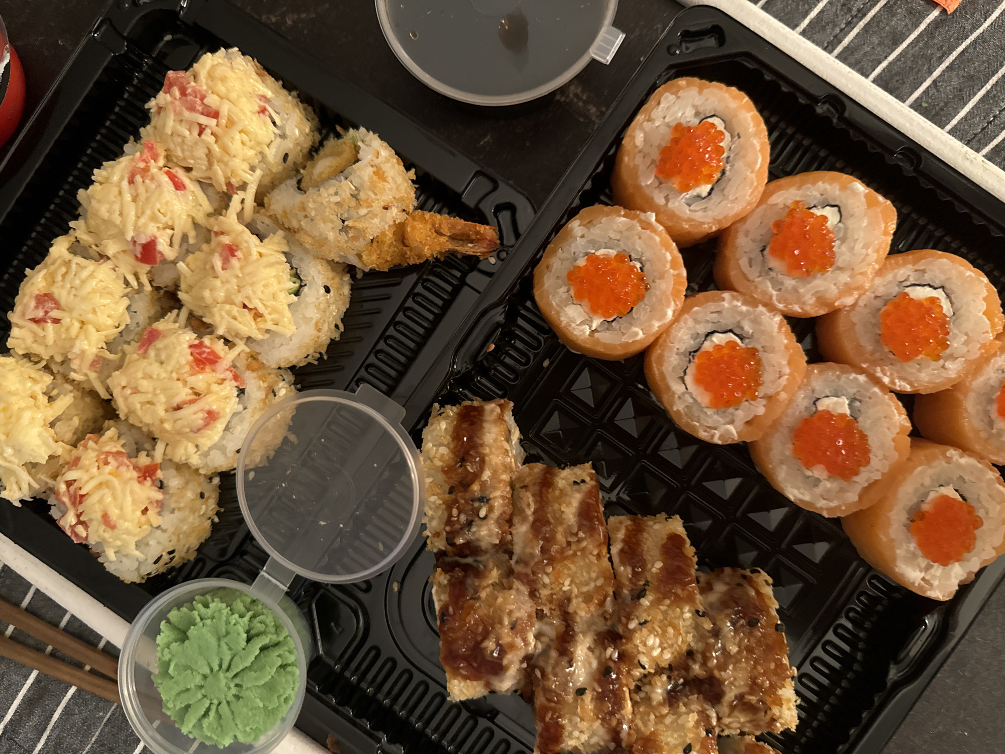 The image shows a black plastic container of sushi. The container is divided into sections containing different types of sushi: one section has sushi topped with shredded imitation crab meat and diced red bell peppers, another has tempura shrimp rolls, another contains salmon rolls topped with salmon roe, and another contains eel rolls. Small containers of soy sauce and wasabi are also visible. Chopsticks are on the side. The setting appears to be a table in a casual dining environment. The exact location is Akulovo (Gp Kubinka), Russia.
