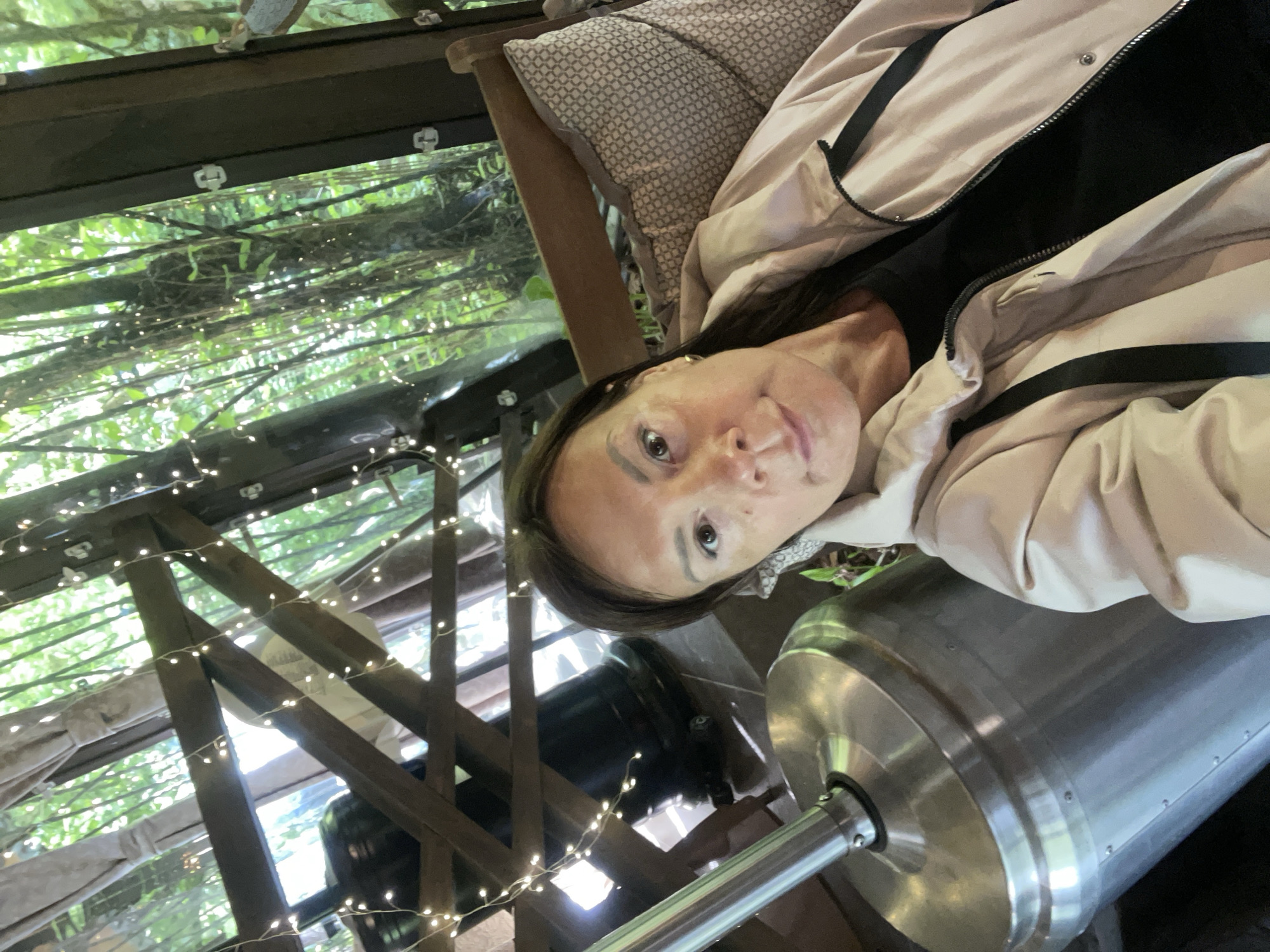 The image is a selfie taken by a woman wearing a light pink jacket over a black top. She is sitting in a chair with a patterned cushion. Behind her, there is a wooden structure adorned with string lights. Through the structure, there is a view of green foliage, possibly trees or plants. To her right, there is a large, cylindrical, stainless steel object, possibly a heater. The location is Solokh-Aul, Russia.