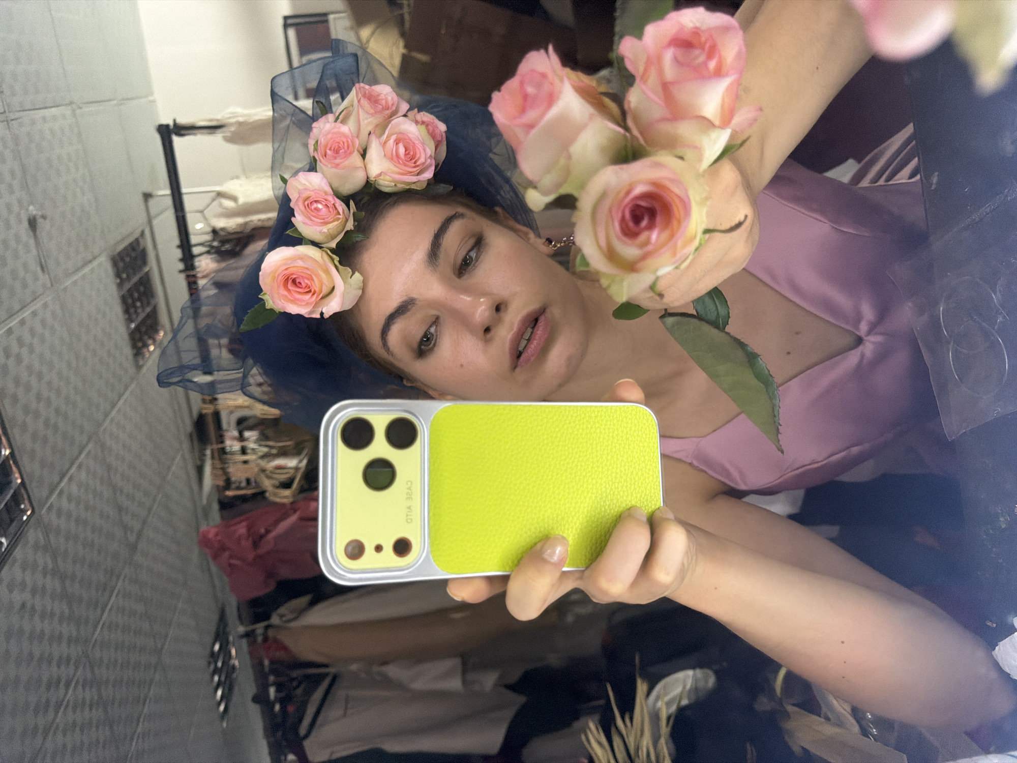 A young woman is taking a selfie in a mirror, holding a neon yellow phone with a silver frame. She is wearing a pink satin dress and a dark blue fascinator adorned with pink roses. She has a bouquet of pink roses in her right hand. The background appears to be a dressing room or backstage area, with clothes hanging on racks. The ceiling is a tiled grid pattern. The woman is looking at the phone camera with her left eye slightly closed and her mouth slightly open. Her expression is contemplative. The lighting is bright and overhead.