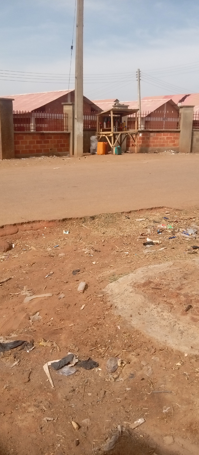 The image displays a daytime street scene in Kasuwan Magani, Nigeria. The foreground consists of a dirt and gravel ground surface extensively littered with various debris, including plastic bottles, bags, paper, and organic matter. This unpaved area borders a slightly elevated, reddish-brown paved road that traverses horizontally across the midground.
In the background, positioned beyond the paved road, a low brick wall topped with a black metal fence delineates a property. Behind this boundary, multiple single-story buildings with red corrugated metal roofs are visible under a clear, light blue sky.
A prominent utility pole, light gray in color, stands vertically on the roadside to the left of the image's center, extending out of frame at the top. Wires are attached to this pole. To its right, near the brick wall, is a small, elevated wooden kiosk with a simple canopy roof. Beneath the kiosk, an orange jerrycan, a green jerrycan, and a grey plastic bag are visible. The top of the kiosk holds numerous smaller containers, consistent with a roadside vending operation. Further to the right in the background, another utility pole with multiple insulators and power lines is present. No individuals or vehicles are depicted in the scene.