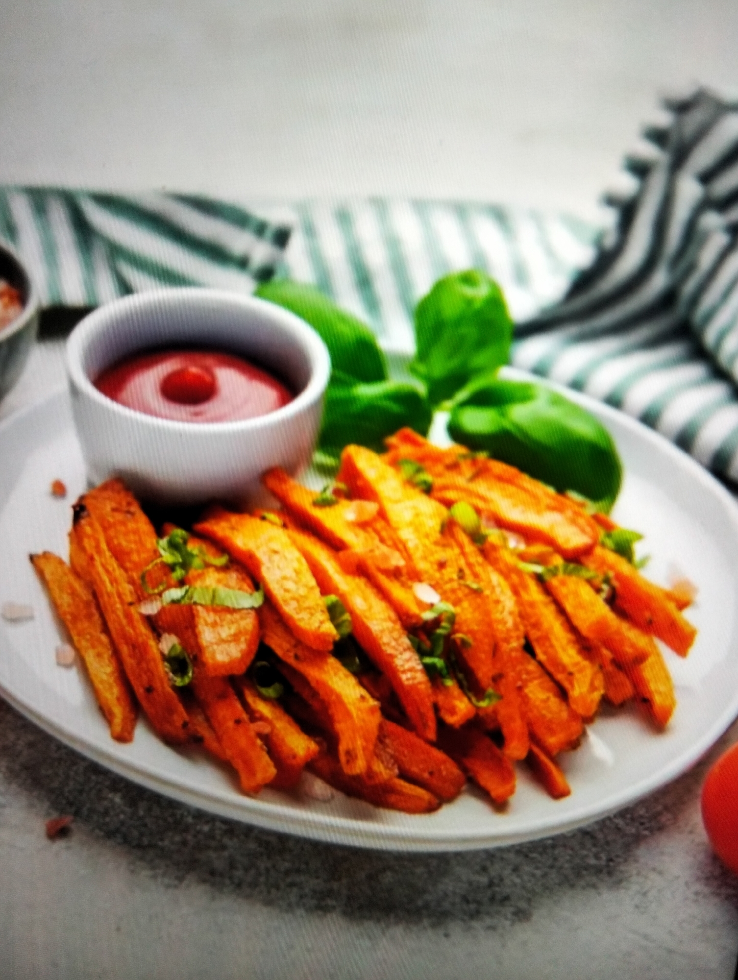 A plate of sweet potato fries is served with a small bowl of ketchup and garnished with chopped green onions and basil leaves. The fries are a vibrant orange color and appear to be seasoned with salt. In the background, a striped green and white napkin is partially visible, along with a few large green basil leaves. The scene is set against a light-colored, textured surface, possibly a countertop or table. The image is a close-up shot, focusing on the food. There is no indication of any specific location in Payagaboh, Indonesia.