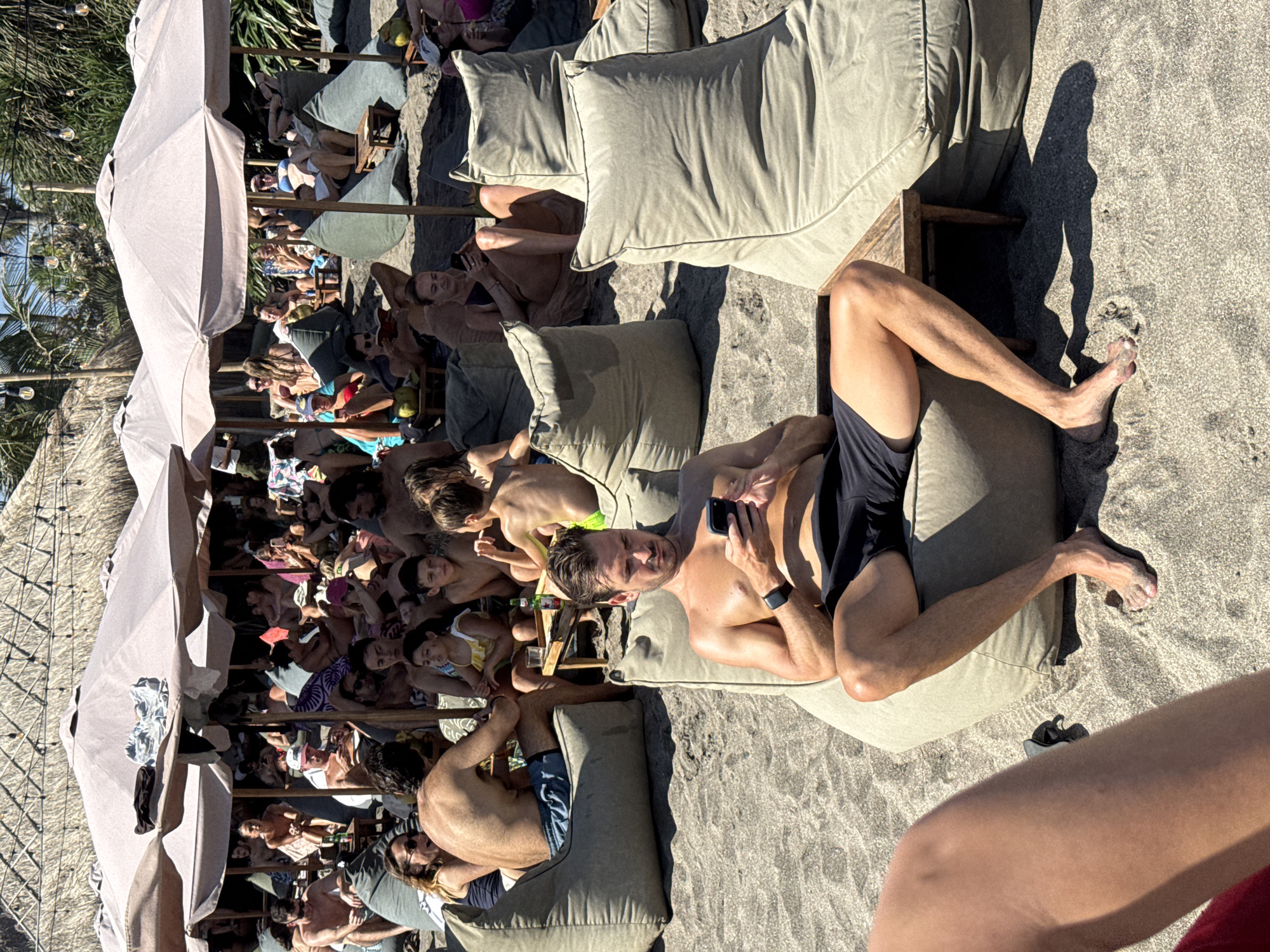 This outdoor photograph captures a crowded scene at a beachside venue in North Kuta, Indonesia. Several people are seated on beige lounge cushions and beanbags arranged on the sandy ground. Umbrellas provide shade to clusters of people, primarily located to the left of the scene.
The foreground focuses on a man lying on his back on a cushion, holding a smartphone, and wearing dark swimwear. Another man lies reclined on a cushion in the lower-left corner of the image, partially visible. In the lower-right corner, there is a partial view of a person's leg.
The environment suggests a tropical beach setting, evident from the sand, umbrellas, and what appears to be thatched roofing above the crowd. The people are engaged in leisure activities, primarily sunbathing and relaxation.