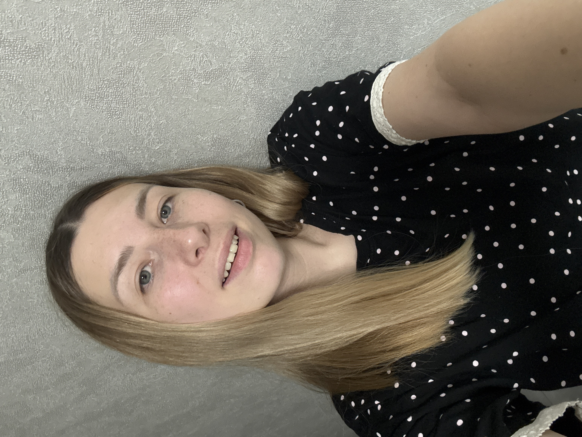 A person with fair skin, light brown hair, and blue eyes is centered in the frame, smiling slightly. The person is wearing a black dress with white polka dots and a white lace trim on the sleeves. The background is a textured gray wall. The image is likely taken indoors. There is no information to determine the location in Moscow, Russia.