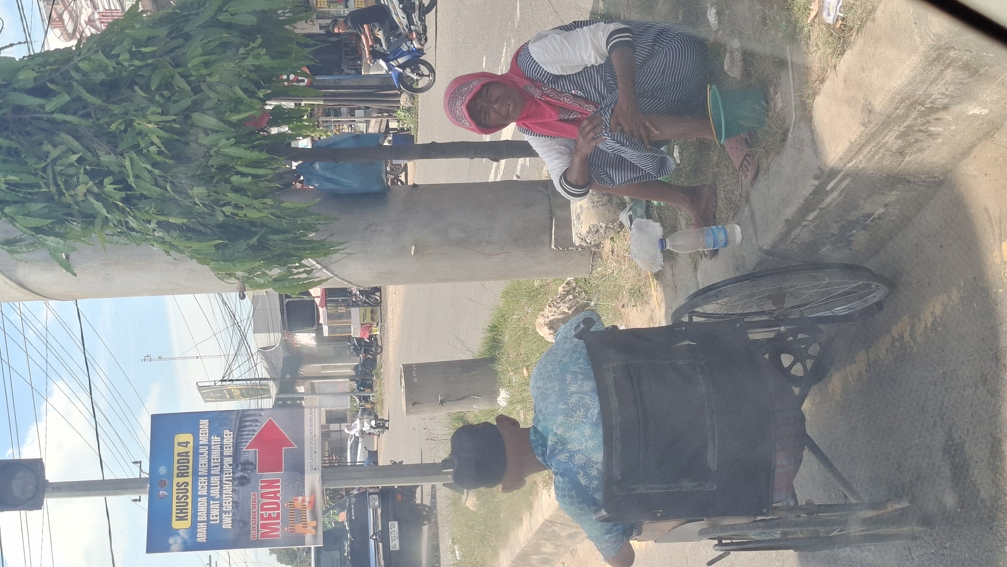 In Pantegajah, Indonesia, a man in a wheelchair faces away from the camera, his back to the street. He wears a blue patterned shirt and a black skullcap. To his right, a woman sits on the curb, facing the viewer. She is wearing a pink headscarf and a striped dress, and her legs are crossed. In front of her is a green bucket. A bottle of water and some trash are also on the ground near the curb. A large tree with green leaves shades the area. In the background, traffic can be seen on the street, including cars and motorcycles. A sign directing traffic to Medan is visible on the left side of the image. The scene appears to be a street corner with a sidewalk and a grassy area.