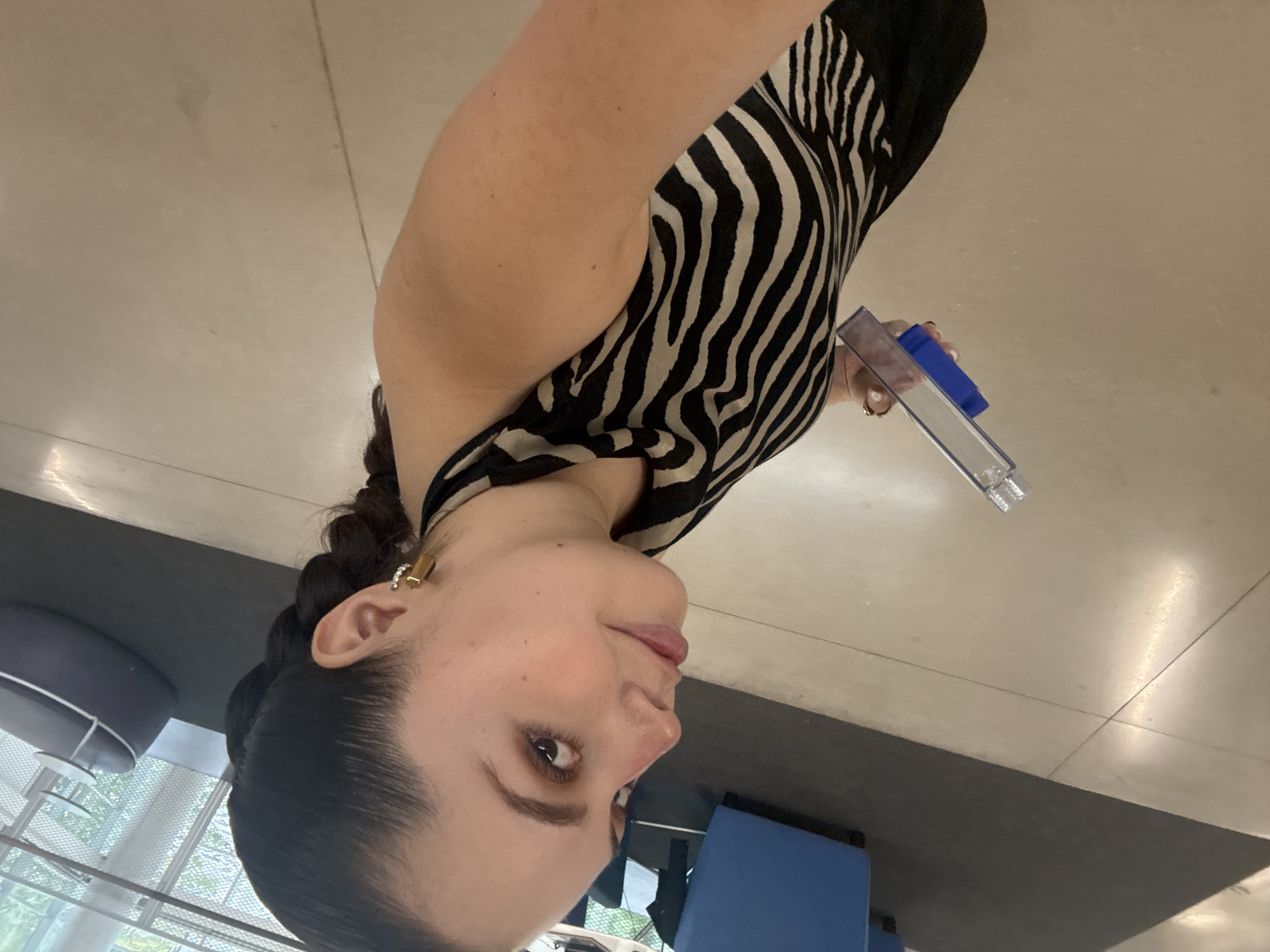 A medium shot depicts a woman indoors. She has dark hair styled in a braid, fair skin, and wears a black and white zebra-striped sleeveless dress. She is holding a clear plastic container with a blue lid. The background includes a beige-tiled floor and indistinct architectural elements suggesting an interior space. Based on the architectural style, the scene might be located in Mexico City, Mexico.