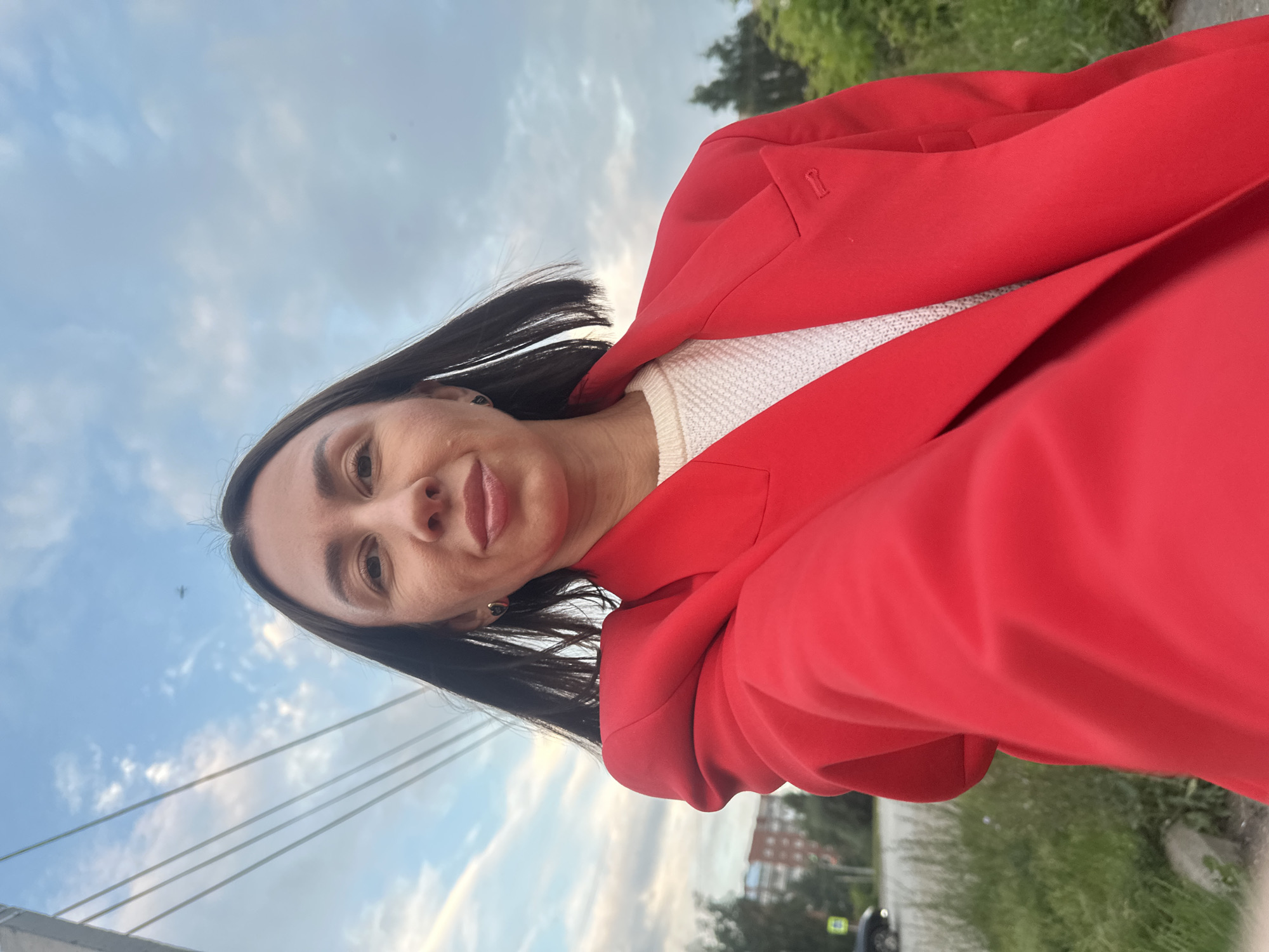 A woman with dark hair, wearing a bright red blazer over a white sweater, is taking a selfie outdoors in Votkinsk, Russia. The background shows a cloudy sky, power lines, some green foliage, and a distant building. A car is visible on a road to the left.
