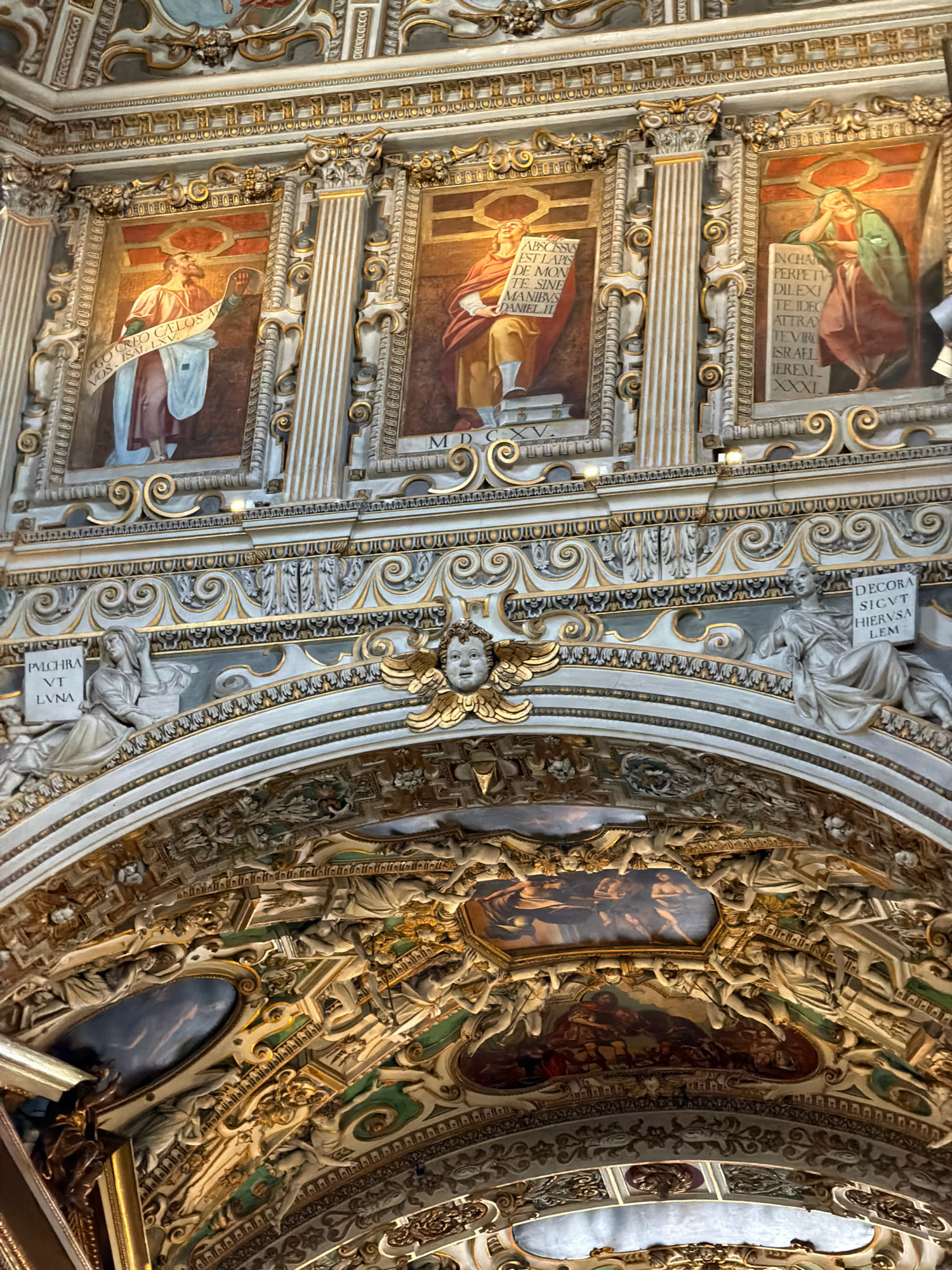 This image captures a section of the ornate ceiling and upper wall of a building, likely a church or cathedral, in Mozzo, Italy. The scene is rich with religious artwork and architectural details, characterized by gilded embellishments, intricate carvings, and painted frescoes.
Dominating the upper portion are three framed paintings, each depicting a male figure with a halo, holding scrolls inscribed with text. The figures are dressed in classical robes, suggesting religious or biblical narratives. Below these paintings, a decorative frieze runs across the width of the image, featuring scrolling patterns and carved figures. On the left, a female figure in a flowing garment is seated, holding a scroll with the visible text "PVLCHRA VT LVNA." To the right, another female figure is also seated, leaning on an armrest, and holding a scroll with the text "DECORA SICVT HIERVSA LEM." Between these figures, a cherubic face with gilded wings is prominently featured.
The lower section of the image reveals a highly elaborate, multi-layered structure of carvings and paintings. These elements include numerous smaller figures, possibly angels or cherubs, and further painted scenes within oval frames, one appearing to depict a biblical event. The overall impression is one of opulence and religious devotion, typical of Baroque or Renaissance ecclesiastical architecture. The lighting suggests an indoor setting, possibly illuminated by artificial lights or natural light filtering from unseen sources. The visible text consists of Latin inscriptions, likely containing scriptural references or dedications.