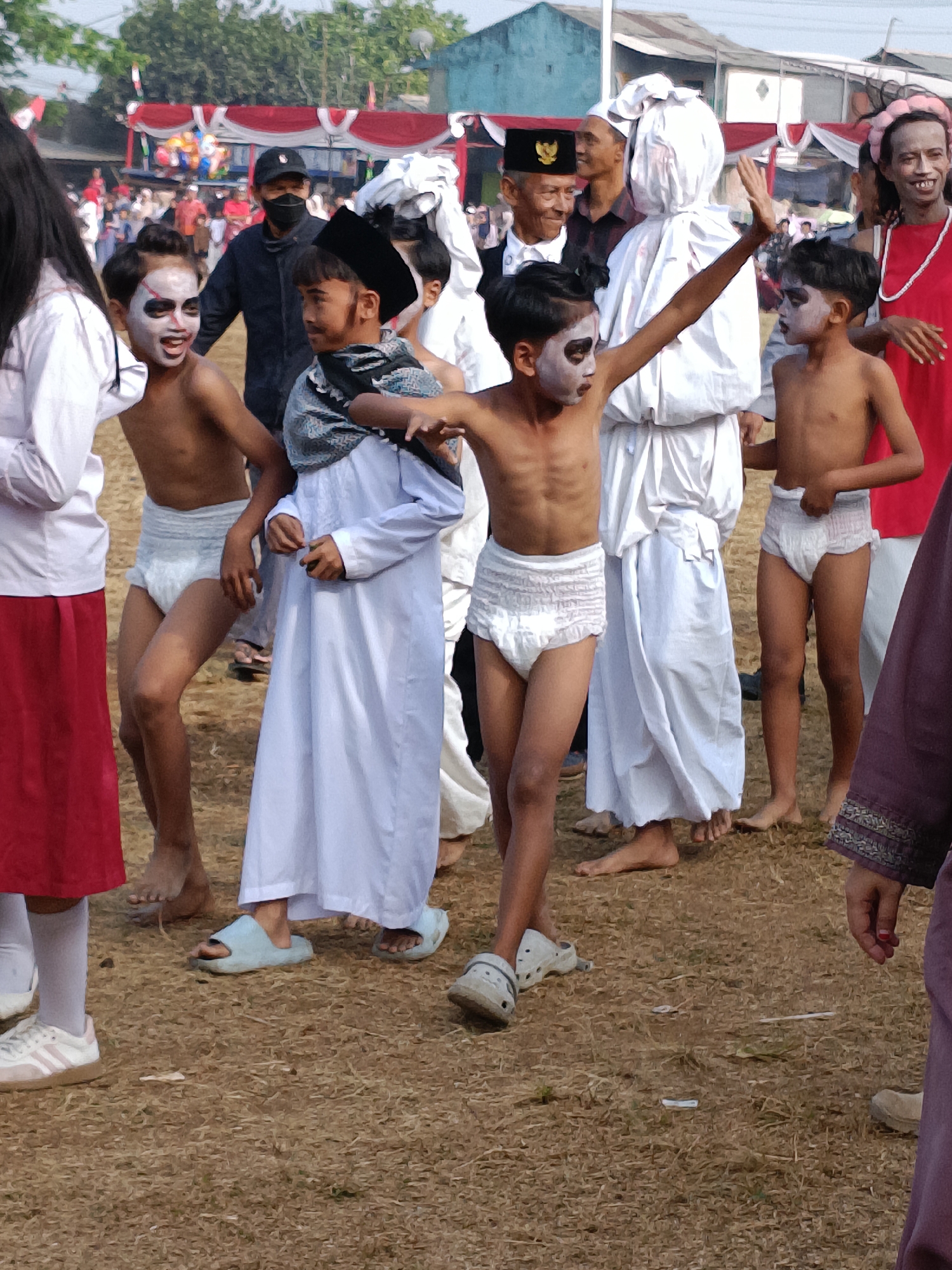 This image depicts an outdoor event in Cibodas, Indonesia, with a group of children and adults gathered on dry, grassy ground. In the foreground, two young boys are the primary focus. The boy on the left, with his mouth open in a yell or laugh, has white and red face paint. He is shirtless and wearing a diaper. To his right, another boy, also with white face paint, is wearing a white Islamic robe, a dark patterned head covering, and a diaper. His right arm is raised in a gesture.
Behind them, other individuals are present. A person in a white shroud-like garment stands between the two boys. To the left of the shrowded figure, a person in a dark jacket and a black face mask is visible. Further back, a crowd of people, some in formal attire with a hat adorned with a national emblem, are observing the event. Red and white banners with balloons are strung up in the background, suggesting a festive or celebratory occasion. The overall scene appears to be a cultural or community gathering.