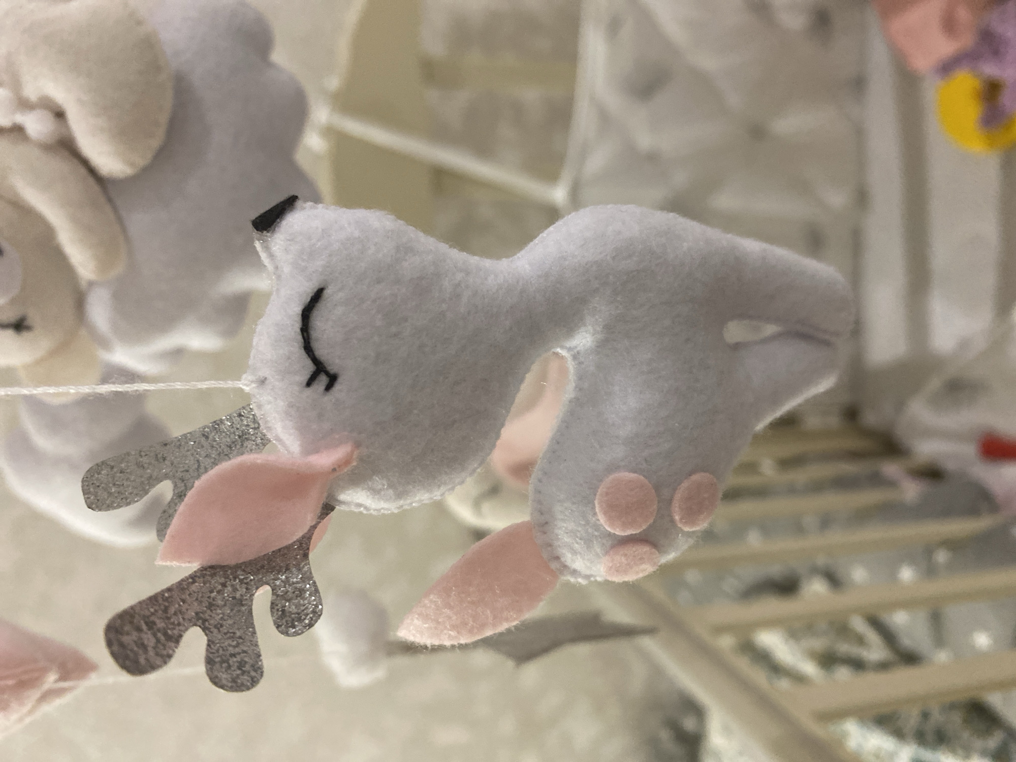 The image depicts a close-up view of a handcrafted baby mobile ornament, specifically a light grey felt deer, hanging in a nursery in Tyumen, Russia. The deer features sparkly silver antlers, soft pink inner ears, a delicate black embroidered closed eye, a small black nose, and a pointed pink felt tail. Three small pink felt circles are affixed to its left hind leg, resembling paw pads. In the background, other elements of the mobile are visible, including a soft white cloud shape with a beige, possibly lamb or bear, figure resting on top. The lower portion of the image reveals parts of a light-colored wooden baby crib with vertical slats and a blurry patterned sheet, grey with white dots. Faint, out-of-focus colorful shapes, including yellow and pink, are discernible in the far bottom background, suggesting other nursery items. The scene is well-lit, providing clear details of the felt toys.