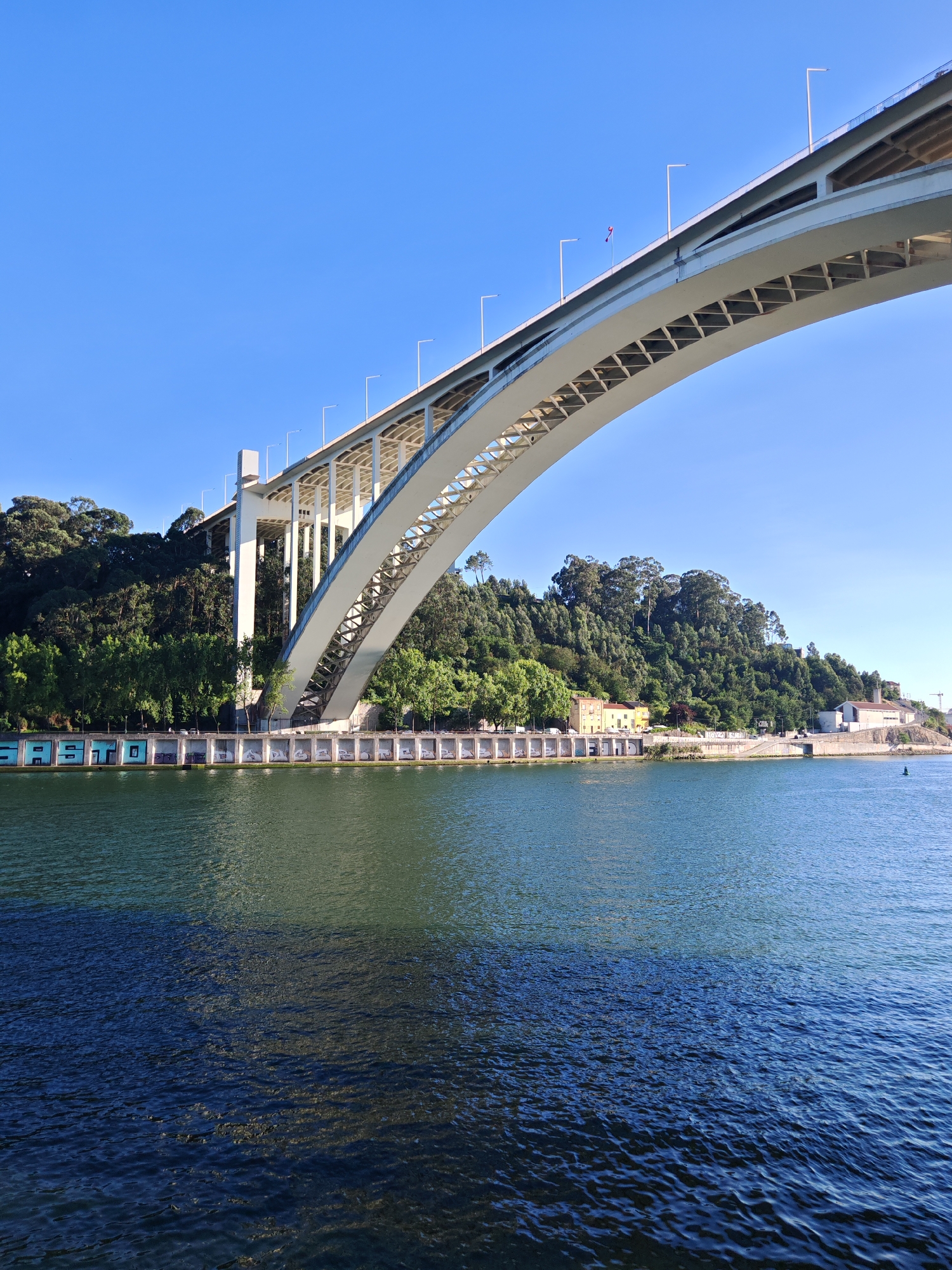 The image shows a wide, arched bridge spanning a body of water under a clear blue sky. The bridge's structure is white and features a latticework design underneath the roadway. Several lampposts are visible along the top of the bridge. On the far bank, a lush, tree-covered hillside rises, with a few buildings nestled at its base. Some of these buildings appear to be historical, with yellow and white facades. Along the waterfront, there is a row of structures, some of which are covered in colorful graffiti. The water is a deep, shimmering green and blue, reflecting the clear sky and the underside of the bridge. The sunlight suggests it is daytime, likely morning or afternoon, with fair weather. The scene evokes a sense of scale and architectural grandeur, with the natural landscape providing a contrasting backdrop. The graffiti on the waterfront buildings adds a touch of urban art to the scene. No people are clearly visible in the image. The text "SASTO" is visible in large, block letters on some of the graffiti-covered structures along the waterfront.