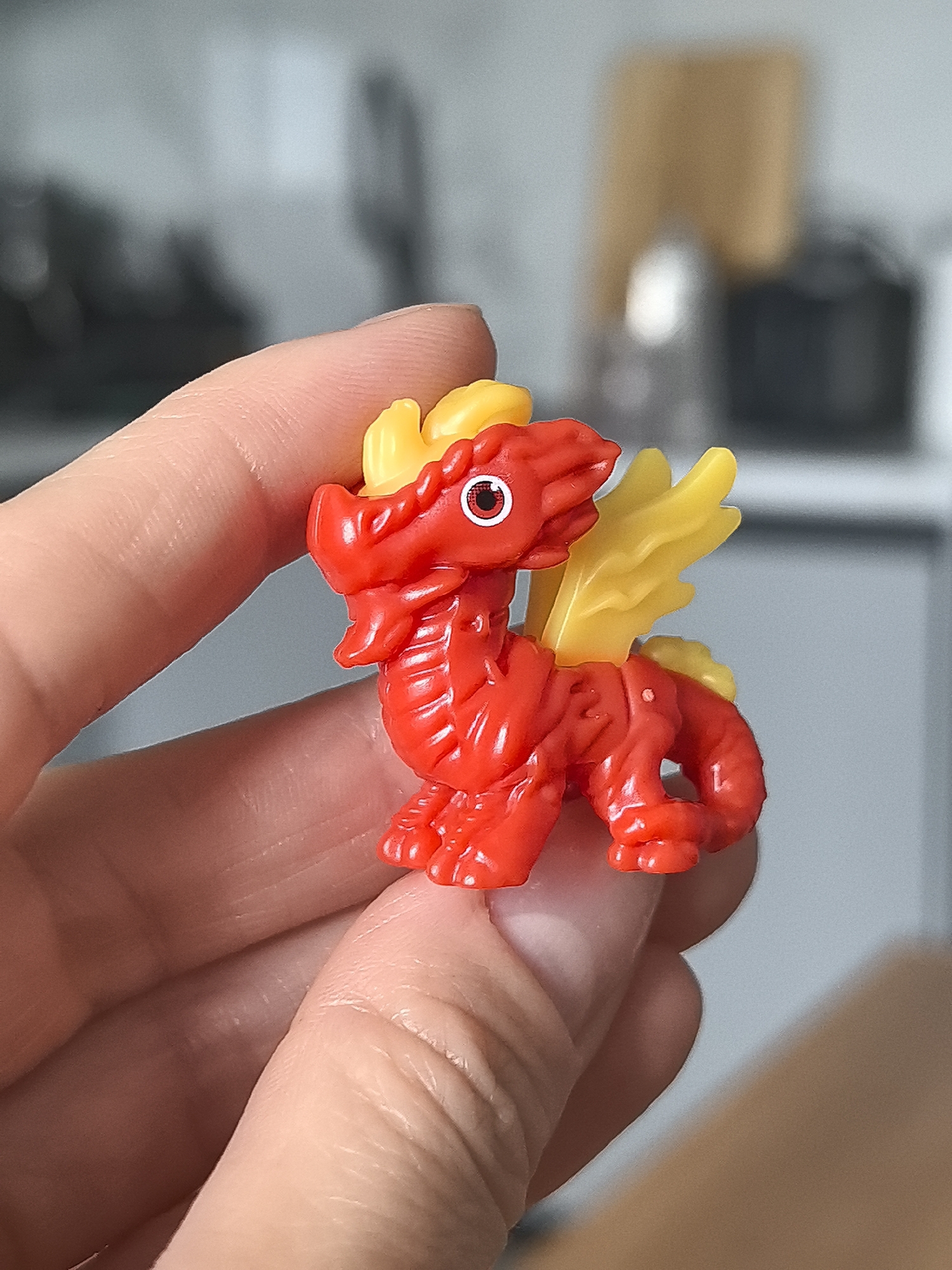 A small, red and yellow plastic dragon toy is held in a person's hand. The dragon has wings and a stylized design. The background is a blurred interior setting, possibly a kitchen or office, with neutral tones. There is no indication of the image being taken in Perm, Russia.