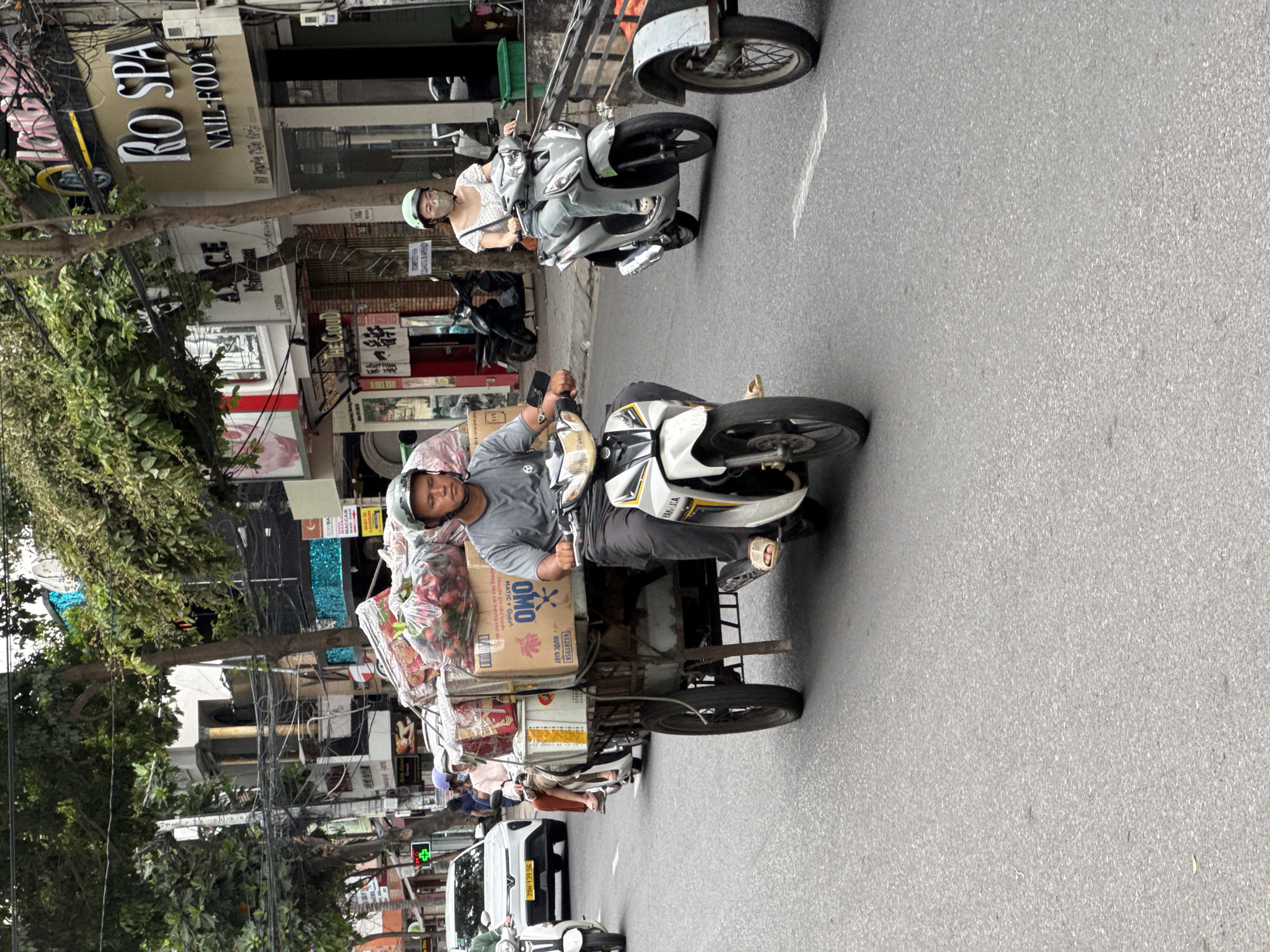 A man wearing a grey t-shirt and a grey helmet rides a white and grey motorbike, pulling a cart filled with goods. The cart is loaded with boxes and large plastic bags containing what appear to be produce, possibly tomatoes or strawberries. To the right of the man, a woman wearing a white top, light blue jeans, and a light green helmet rides a silver motorbike. The street is paved and appears to be a city street in Nha Trang, Vietnam. Buildings line the street, some with storefronts. Traffic is light with a white car visible in the distance. The overall scene depicts daily life and commerce on a busy street in Vietnam.