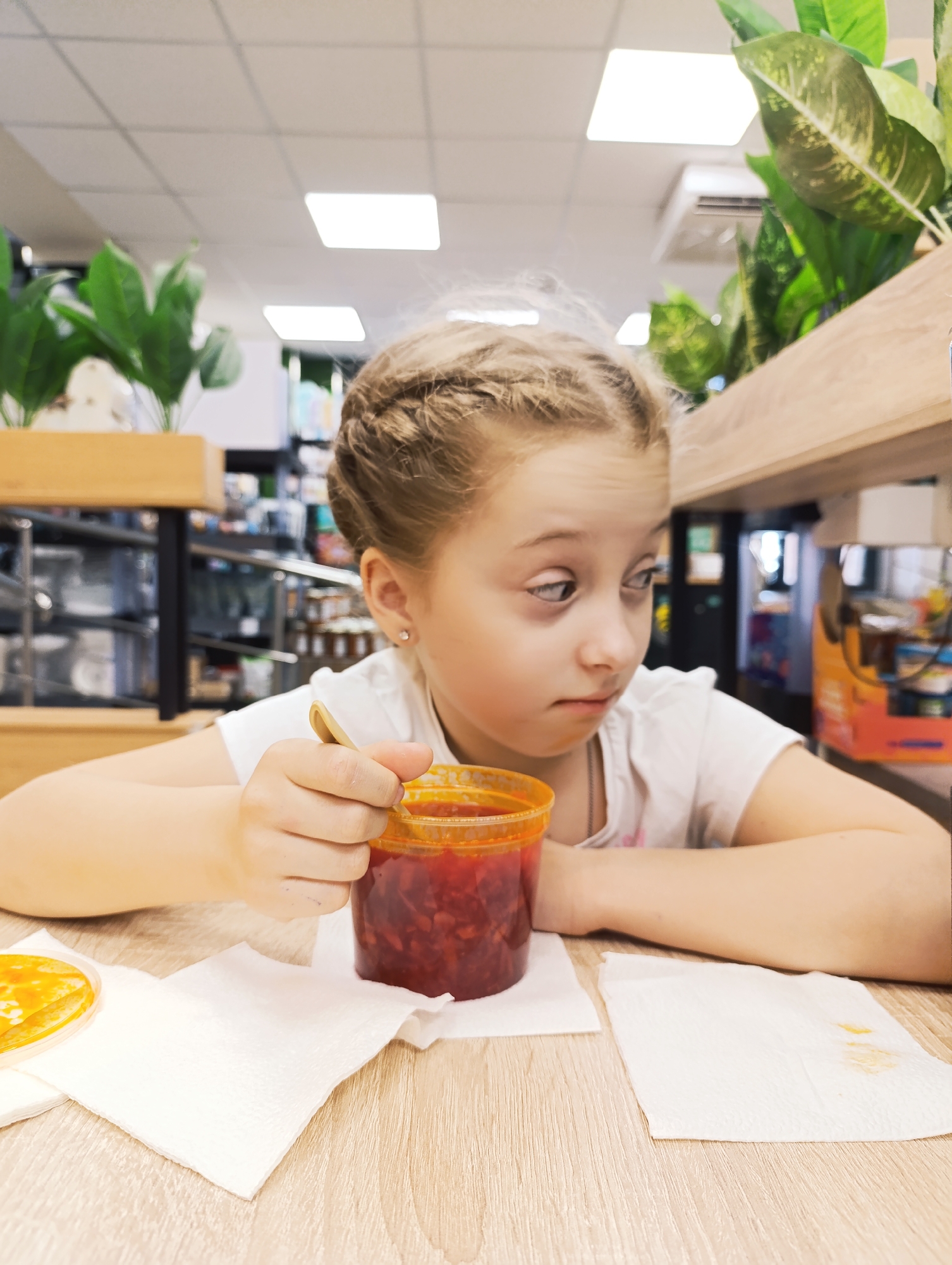 A young girl with braided blonde hair is seated at a light-colored wooden table, holding a plastic container filled with a red, soup-like substance. She is holding a spoon in her right hand and gazing to the right with a somewhat skeptical expression. Two white napkins are on the table around her. The girl is wearing a white T-shirt.
Behind her, the background includes indoor plants in wooden planters, shelves with various items, and a ceiling with recessed lighting. An air conditioning unit is visible near the ceiling. The scene appears to be indoors, possibly in a cafeteria or a restaurant.
The location is in an unknown city, in an unknown country.
