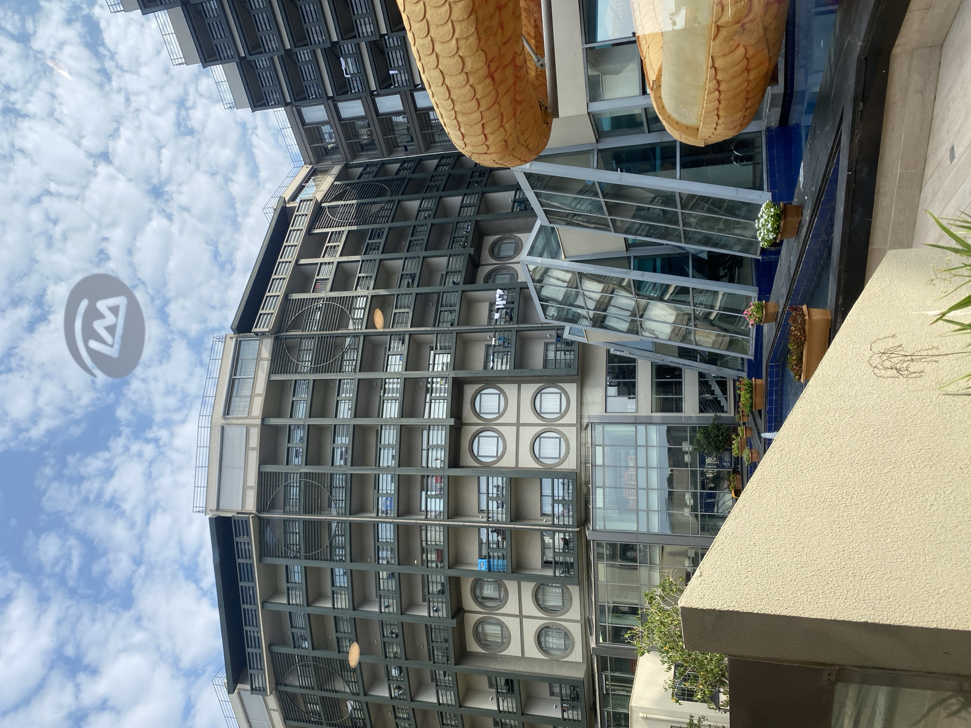 A wide-angle, eye-level outdoor shot captures a large, modern hotel building complex under a cloudy sky. The building, possibly in Aksu, Turkey, features a curved facade with numerous balconies and circular windows. A contemporary architectural style is evident, with glass elements and tiered structures. In the foreground, a light-colored, textured wall forms a triangular shape. To the right, a large, gold, scaly sculpture, resembling a dragon or snake, curves into the frame. The scene suggests a resort or hotel environment with a pool area visible in the background, indicated by blue tiling and planters with flowers. The overall atmosphere is bright, with diffused sunlight filtering through the clouds.