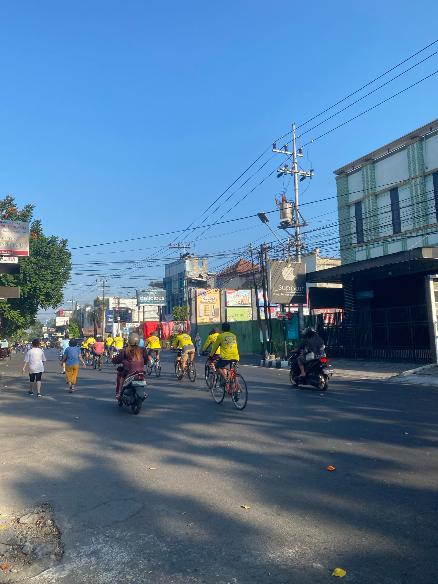 The image captures a bustling street scene in Kediri City, Indonesia, under a clear blue sky. A group of people are engaged in various activities on the road. Several individuals are cycling, with many wearing bright yellow shirts. Others are walking, and one person is riding a scooter. The road is paved and shows signs of wear, with some debris on the side. Buildings line the street, featuring advertisements and signs, indicating a commercial area. Electrical poles and wires are prominent fixtures, suggesting a well-developed urban environment. The lighting suggests it is daytime, likely morning or afternoon, given the shadows cast by the sun. The overall atmosphere is one of community activity and daily life in the city.