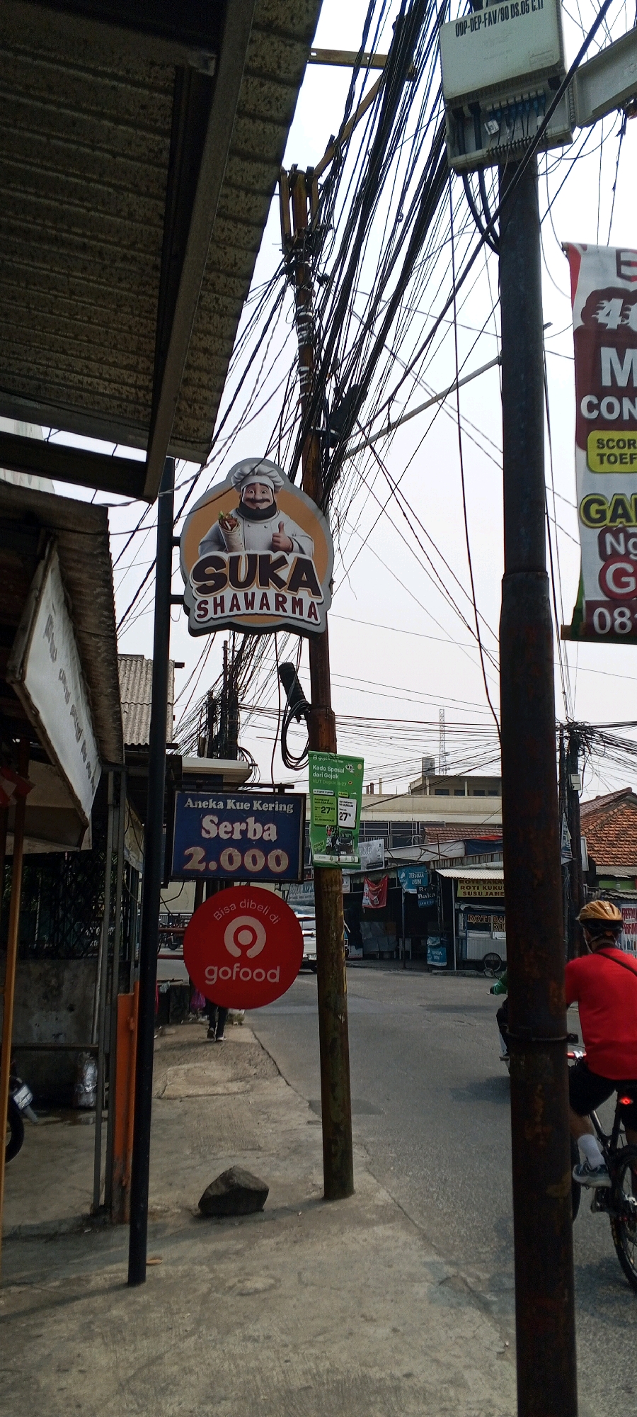 The image depicts a street scene in Depok, Indonesia, characterized by a dense network of overhead electrical wires. A prominent sign for "SUKA SHWARMA" featuring a chef illustration is mounted on a pole. Below it, another sign advertises "Aneka Kue Kering Serba 2.000" (Assorted Dry Cakes All for 2,000) and a red circular GoFood logo. A person is walking on the sidewalk past these signs. In the background, buildings and street infrastructure are visible, along with a person on a bicycle in the foreground, on the right side of the image. The environment appears to be a commercial or mixed-use area with clear skies.