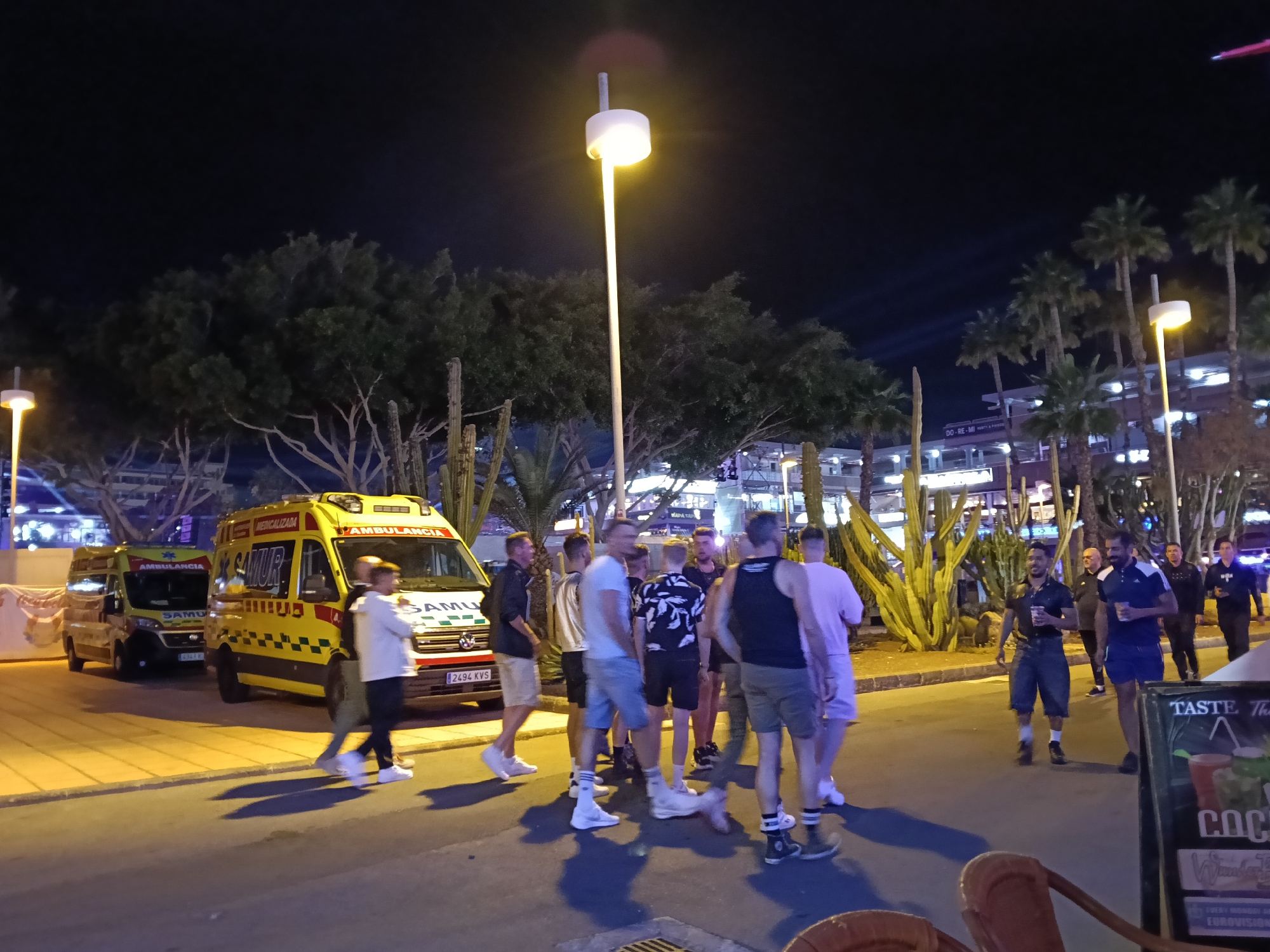 The image depicts a nighttime scene in San Bartolomé de Tirajana, Spain, featuring a group of approximately 15 people gathered on a street illuminated by streetlights. Two ambulances, marked "SAMU" and "AMBULANCIA," are parked on the side of the road. Palm trees and cactus plants are visible in the background, indicating a tropical or arid environment. Some individuals in the group are holding cups or glasses. A bar sign advertising "COCKTAILS" is partially visible. The setting appears to be a tourist or entertainment area due to the presence of bars and the casual attire of the people.
