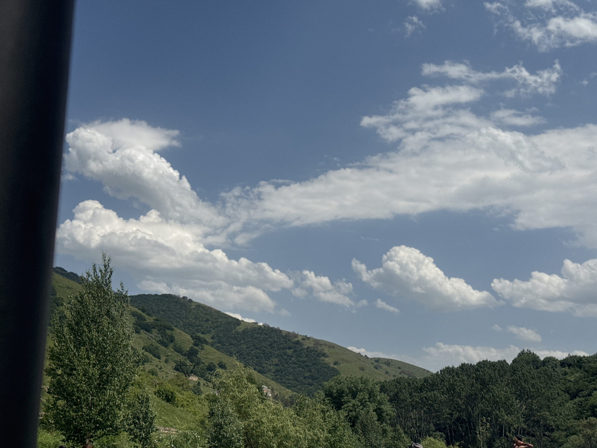 The image depicts a mountainous landscape under a partly cloudy blue sky. The foreground is dominated by lush green vegetation, with a large tree on the left side. In the midground, rolling hills covered in dense forests ascend towards the background. The sky is dotted with fluffy white cumulus clouds. This scene is likely located in Aghveran, Armenia, known for its mountainous terrain and natural beauty, popular for outdoor recreation and as a resort area.