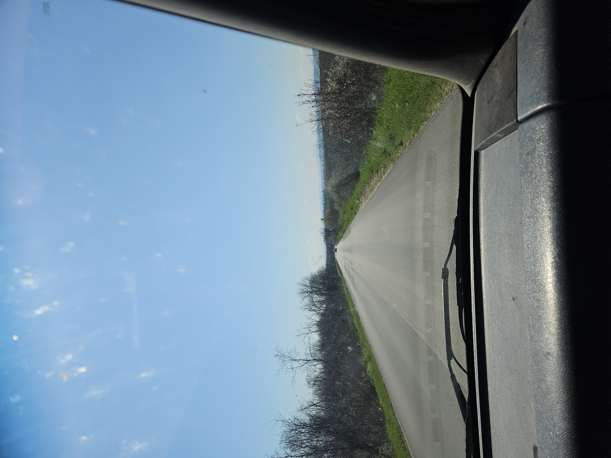 The image shows a view from inside a vehicle, likely a car, driving along a straight, paved road. The road is flanked by vegetation, including trees and grass. The sky is clear and blue with some wispy clouds. The vehicle's windshield and a portion of the interior are visible, including a windshield wiper. The location is likely in the countryside, possibly near Preslavets, Bulgaria, given the presence of a straight road and surrounding vegetation. There is a car visible at the end of the straight line of the road. The picture is rotated vertically.