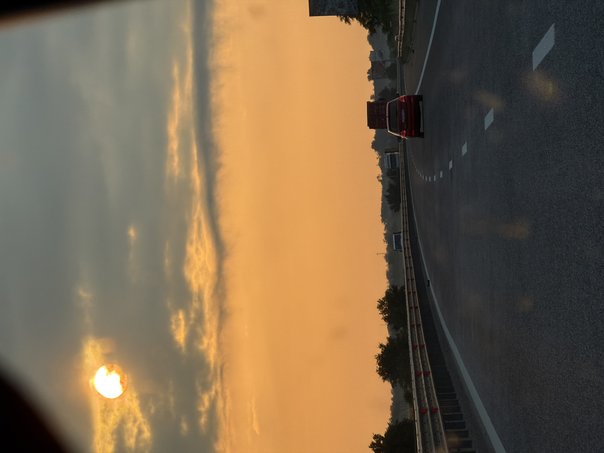 The image, rotated 90 degrees clockwise from its natural orientation, captures a multi-lane highway scene during either sunrise or sunset. The most prominent feature is the low sun, a bright, glowing orb, positioned on the left side of the frame. It bathes the sky in a vibrant gradient of orange and yellow near the horizon, fading into soft grey-blue clouds higher up. The warm, diffused light suggests either dawn or dusk.
The highway, appearing on the right side of the frame, stretches into the distance. It features dark asphalt pavement with distinct white dashed lane markings and a solid white line delineating the shoulder. A metal crash barrier, equipped with red reflectors, runs along the right edge of the road, separating it from the surrounding landscape of trees and vegetation. In the background, beyond the trees, faint outlines of buildings or structures are visible.
Several vehicles are present on the highway. A red passenger car, possibly a hatchback, is seen in the middle ground, heading in the same direction as the viewer. Further down the road, other vehicles, including a large truck or container, are visible. No individuals are discernible in the image. The atmospheric conditions, with the low sun and rich sky colors, evoke a calm and picturesque travel experience. The provided context places the location in Rupkite, Bulgaria, indicating a European highway setting. No text is clearly readable on any objects, including the license plates of the vehicles.