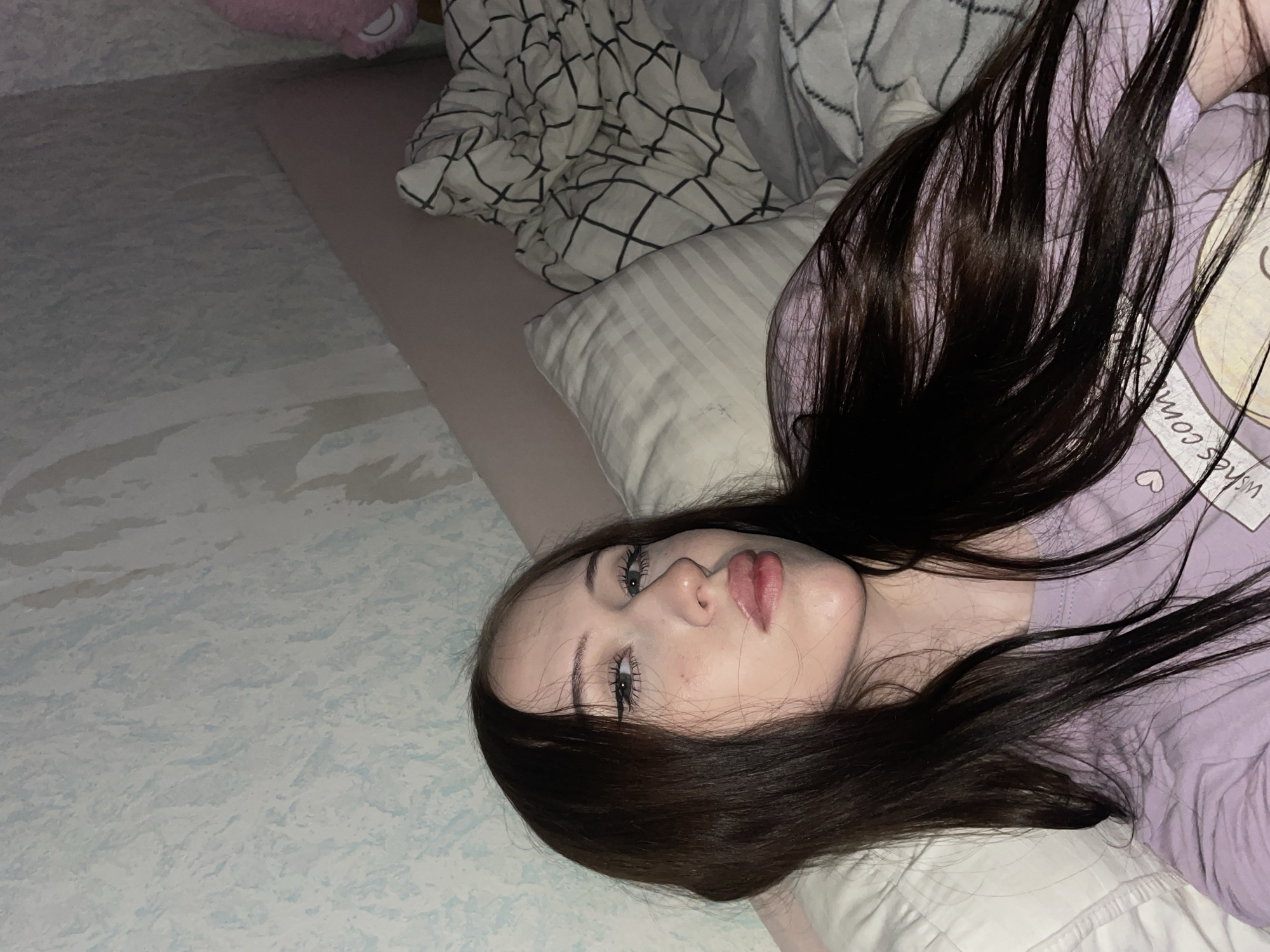 The image shows a young woman lying on a bed, with her head resting on a pillow. She has long, dark hair that is spread around her. She is wearing a light purple t-shirt with some cartoon-like graphics and text. The room appears to be dimly lit, suggesting it is either nighttime or the curtains are drawn. The bedding includes a striped pillow and a duvet with a black and white grid pattern. The overall mood is relaxed and intimate. There is some text visible on the t-shirt, but it is partially obscured. The scene is set in a bedroom, likely in Votkinsk, Russia, given the location context.