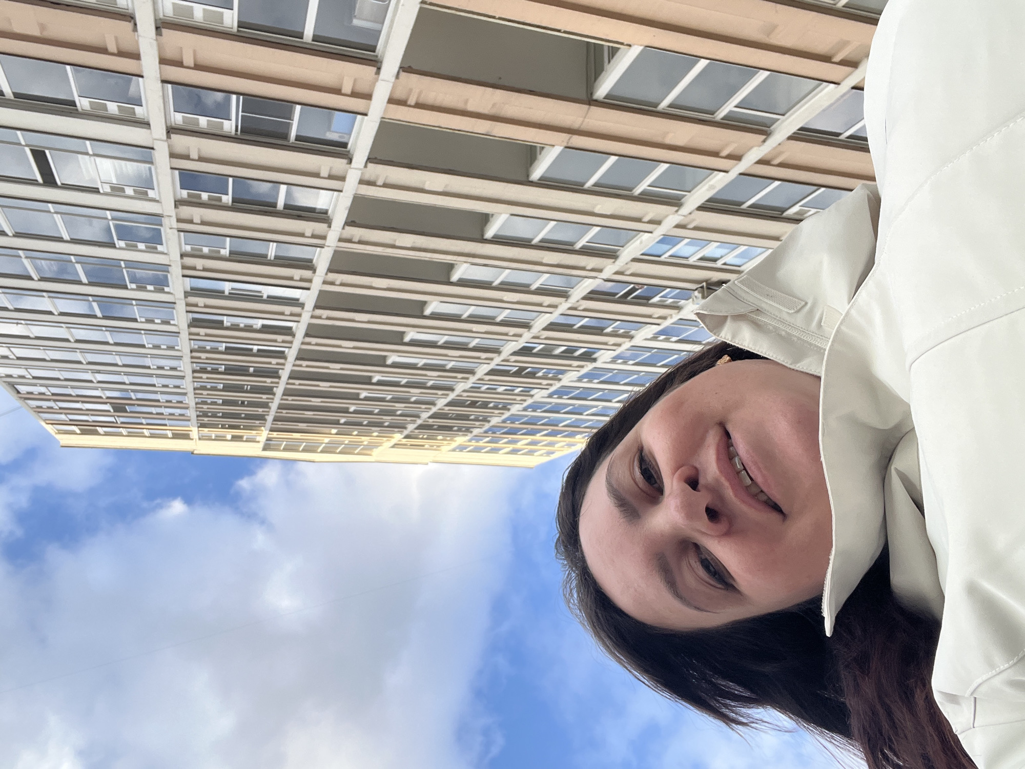 A young woman is depicted in a low-angle, close-up shot, smiling softly at the camera with a pleasant, relaxed expression. She has dark hair and is wearing a light-colored, possibly off-white or very light beige, jacket with visible seams and a collar. Her face occupies the lower right portion of the frame (when viewed with the person upright).
The background is dominated by a clear blue sky with scattered white, fluffy clouds, suggesting fair weather during the daytime. A tall, multi-story building, likely an apartment or office complex, rises vertically in the upper and central background. Its facade features a repeating grid pattern of numerous rectangular windows, many reflecting the sky and clouds. The building's structure includes horizontal bands and vertical elements in tones of light tan or beige and darker gray, creating a uniform and repetitive design.
The image conveys a casual outdoor moment, potentially a selfie taken while looking up at the surrounding architecture and sky in Perm, Russia. No visible text is present.