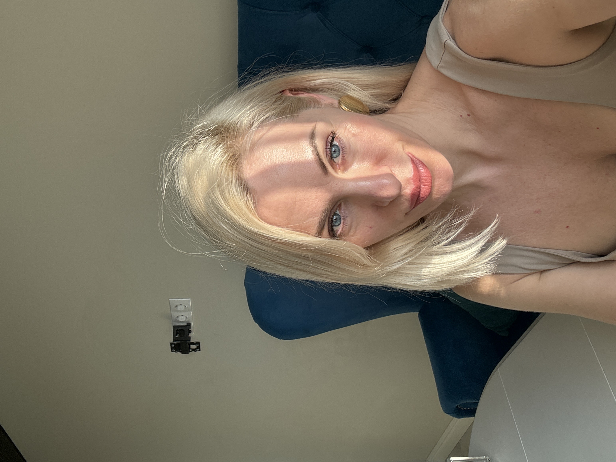 A woman with blonde hair and blue eyes is reclining on a blue chair. She is wearing a beige top. Behind her is a light-colored wall with an electrical outlet. To her right is a white table or surface. The location is Unknown, Unknown.
