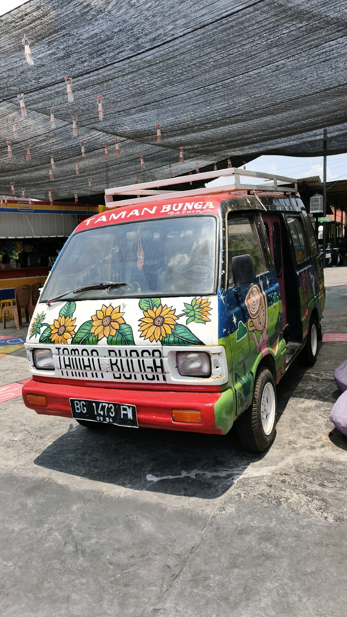 A colorful Suzuki Carry van, brightly painted with sunflowers and a whimsical cartoon character, is parked outdoors on a paved surface. The van's front is white with large, stylized sunflowers and green leaves. Above the windshield, the red text "TAMAN BUNGA" and "Baturaja" is visible. The side of the van features a green and blue mural of a starry night sky with a cartoon character holding a steaming cup. A white roof rack is mounted on top. The van's license plate reads "BG 1473 FW". The ground is a gray, textured concrete with shadows cast by the van and overhead structures. In the background, a black mesh shade structure is suspended by wires, with small, dangling white and red decorative items. Other elements in the background include yellow chairs, a building with colorful signage, and glimpses of other vehicles. The overall scene suggests an outdoor establishment or market in a tropical or sub-tropical location, likely a roadside attraction or a business catering to tourists. The mention of "Baturaja" indicates the specific location within Air Paoh, Indonesia.