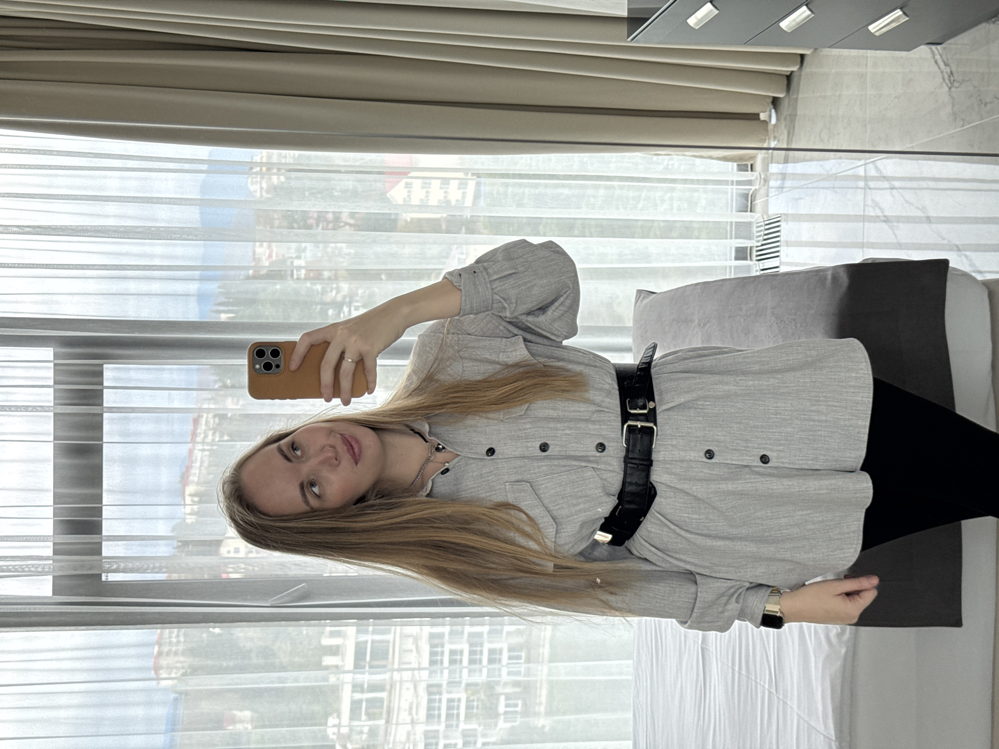 A woman is taking a selfie in front of a window in Sochi, Russia. She is wearing a gray button-down shirt with a black belt and black pants. The window is partially covered by sheer curtains, revealing a view of buildings and the sea. There is a bed behind her with gray and dark gray pillows.