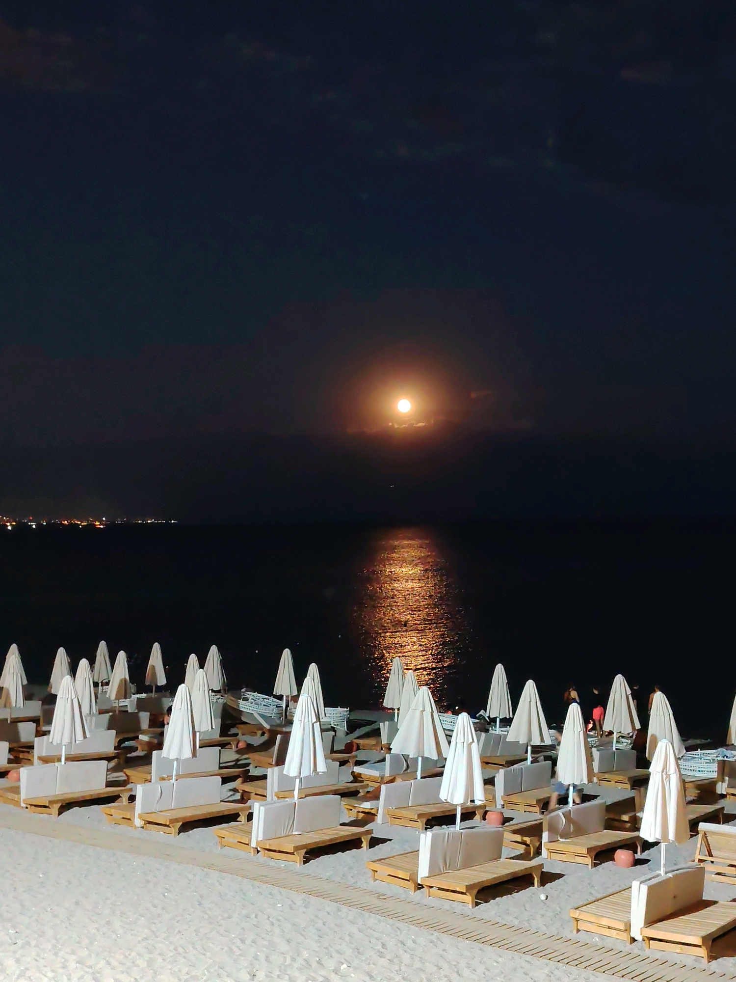 The image depicts a nighttime scene on a beach in Konyaaltı, Turkey, featuring rows of empty lounge chairs and closed beach umbrellas arranged on the sand. The sea is visible in the background, reflecting the light of a large, bright moon. A distant city skyline with illuminated buildings can be seen along the coast. A few individuals are present near the water's edge, some appearing to be standing or walking. The overall atmosphere is tranquil and serene, with the moonlight casting a soft glow over the scene.