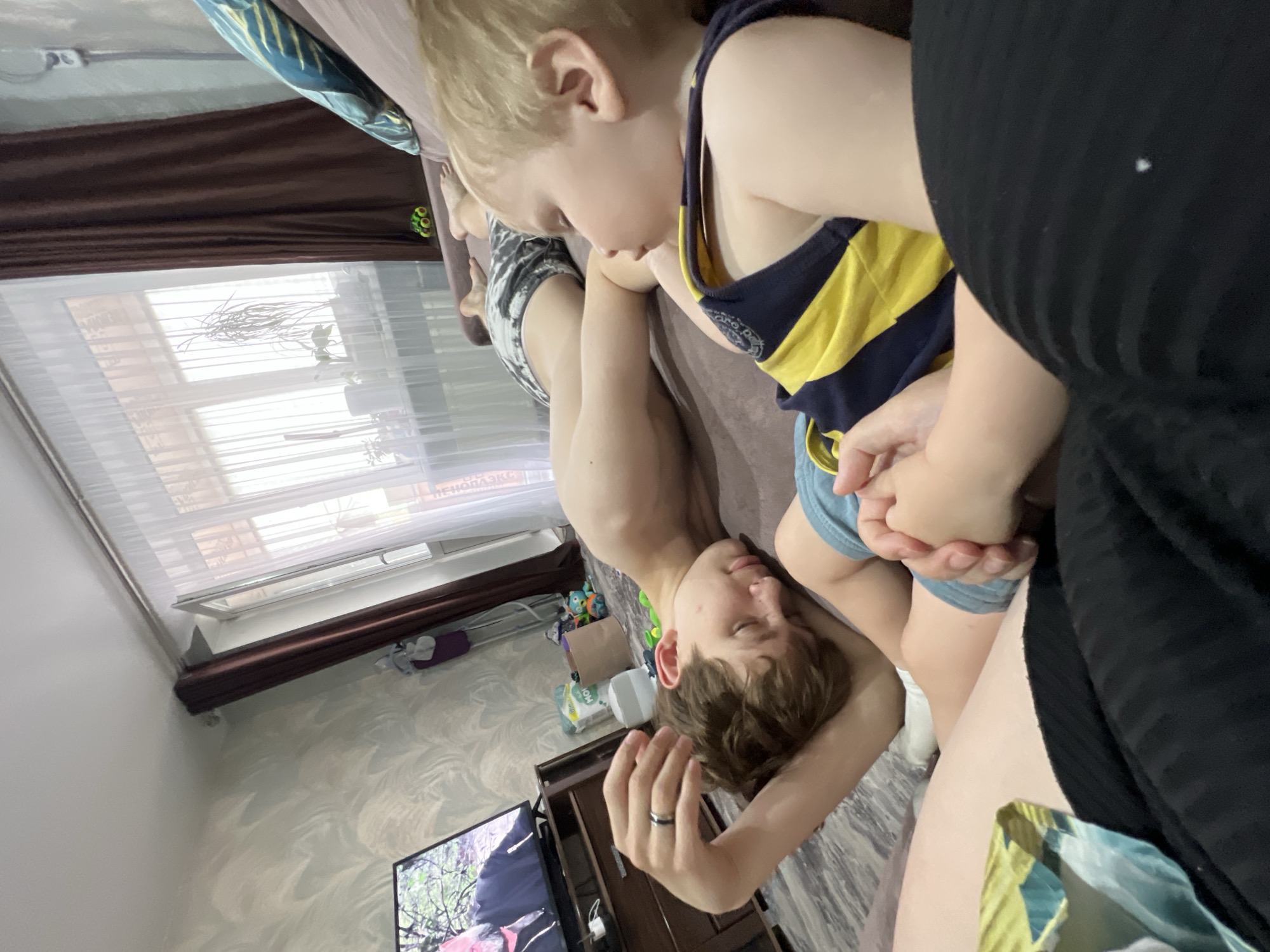 A photo depicts a child in a blue and yellow striped shirt seated on the lap of a person with dark hair and fair skin. The person has their head resting on the child's lap, with an arm raised and a ring visible on one finger. The background shows a room with a window covered by sheer blinds, with brown curtains above. A television screen is visible, along with other assorted objects such as cleaning supplies. The backdrop includes a partially obscured inscription in Cyrillic script, resembling "ПЕНОПЛАЗ", possibly referencing the city of Noviy.