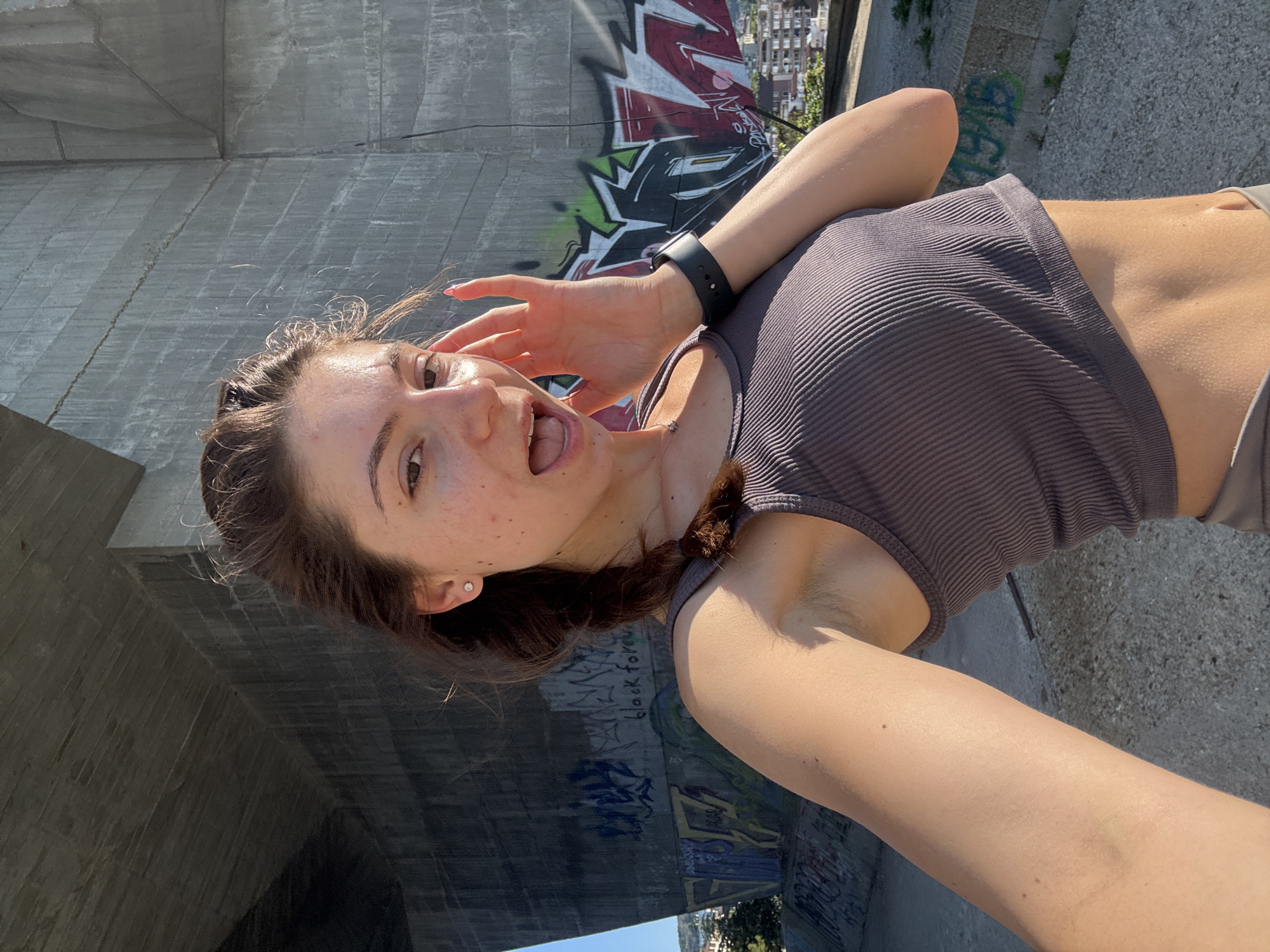 A young woman with brown hair in a braid and a grey ribbed crop top stands in front of a concrete structure with graffiti. Her left arm is extended towards the camera, and her right hand is touching her face. Sunlight casts shadows across the scene. The background reveals a cityscape with buildings and vegetation, suggesting an urban outdoor setting, likely Varna, Bulgaria, given the provided context.