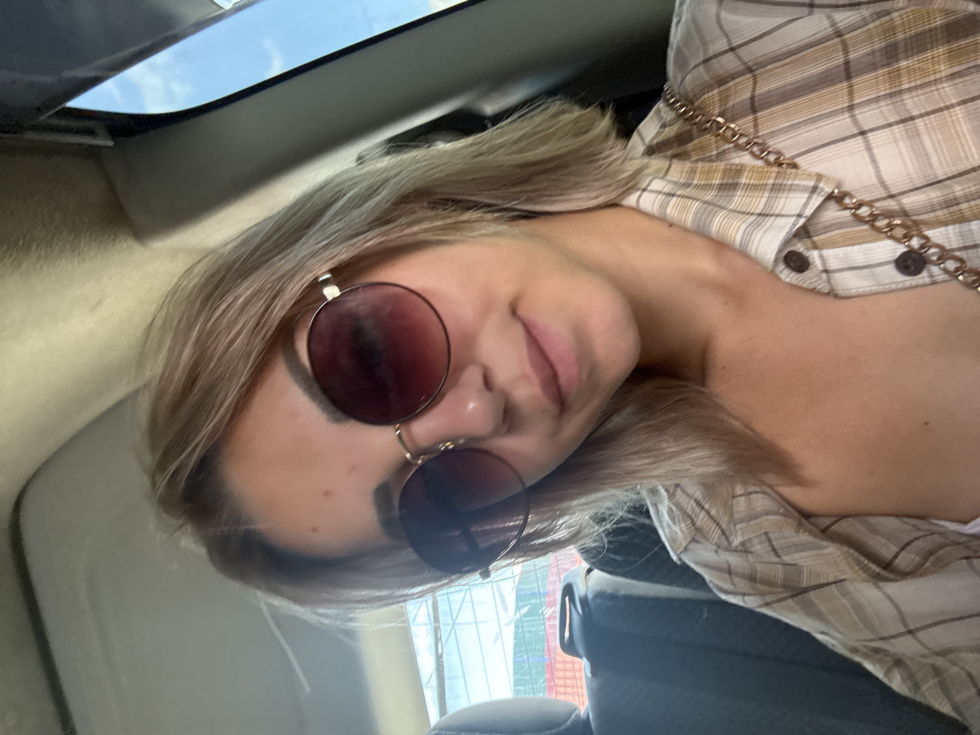 A young Caucasian woman with blonde hair and sunglasses is sitting in the passenger seat of a vehicle. She is wearing a plaid shirt and a gold chain necklace. The background shows the interior of the car, including a headrest and part of the dashboard. Outside the window, a blurred view of buildings and trees is visible, suggesting an urban environment. The lighting indicates it is daytime.