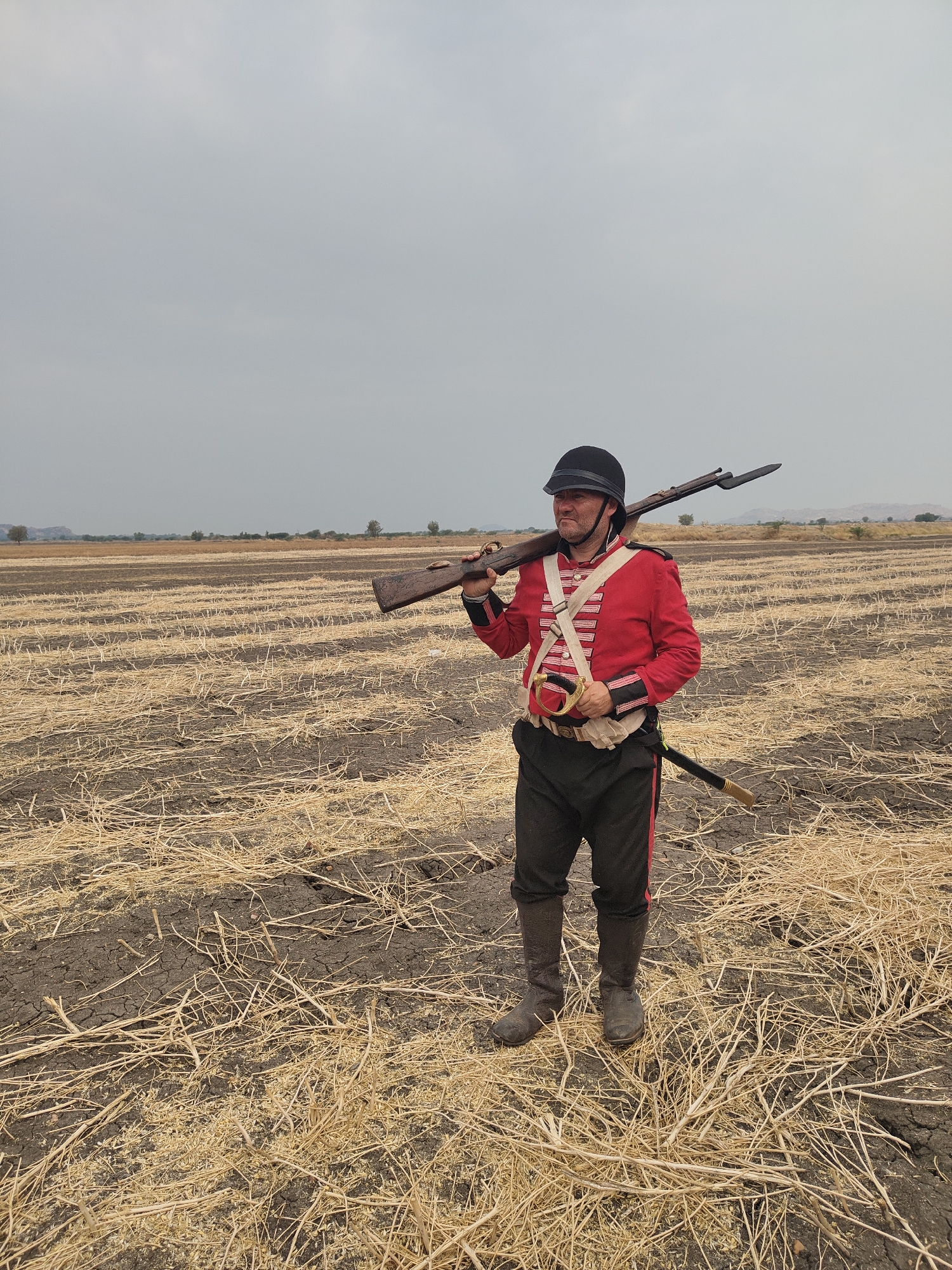 A male individual stands centered in a harvested field in Kottalapalli, India. He wears historical military attire: a black helmet, a red jacket with white cross belts, dark trousers with a red side stripe, and dark boots. A long, dark rifle with an attached bayonet is carried over his left shoulder, held by his right hand. A sabre in a black scabbard with a golden hilt is visible at his left hip, and a light-colored pouch is at his right hip. The man has a slight beard and looks towards the left side of the frame. The field features dark, cracked soil with linear arrangements of dry, golden-brown stubble. The background consists of distant low hills and sparse trees under an overcast, light grey sky.