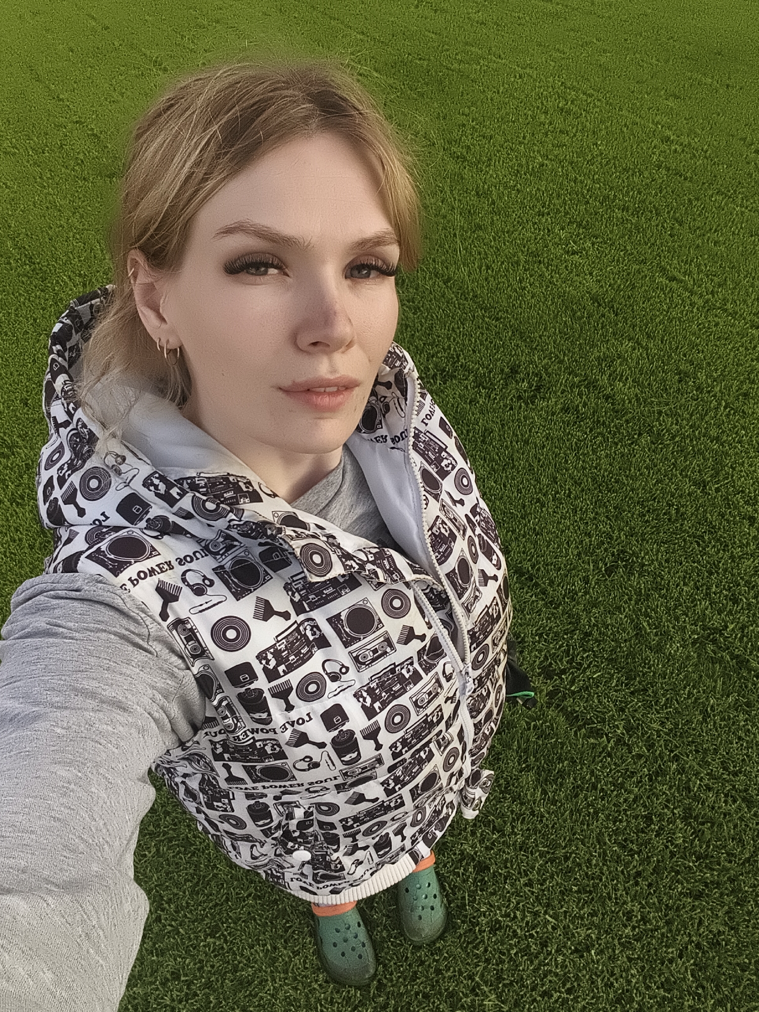 A young woman is captured in a high-angle, full-body selfie, standing on a uniformly green surface that appears to be artificial turf or a very well-maintained natural lawn. The setting is outdoors in bright daylight, indicating clear weather.
The woman has light skin, light brown or blonde hair pulled back, and noticeable dark eyelashes. She looks directly at the camera with a neutral expression. She is wearing a grey long-sleeved shirt layered under a white puffer vest. The vest features a repetitive black pattern depicting various retro audio devices, including boomboxes, vinyl records, and headphones, along with other abstract shapes and what look like brushes. Some text elements are visible within the pattern on the vest, appearing mirrored, such as "BOME X 200J".
Her feet are visible at the bottom of the frame, clad in bright orange socks and green clogs. The background consists solely of the dense, short green ground, which stretches out behind and to the sides of her. The location context is Русский Вожой, Russia.
