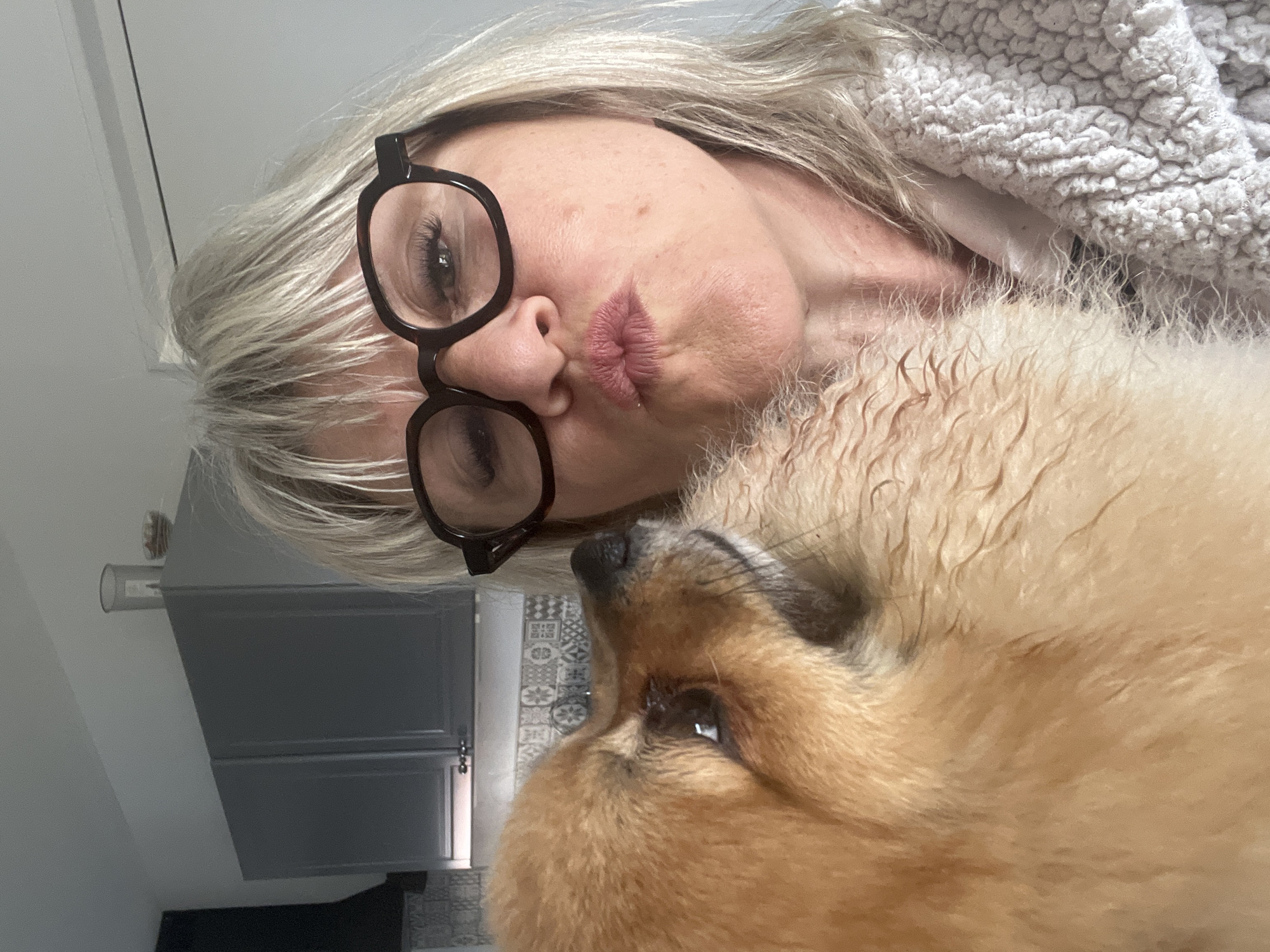 An indoor shot taken in Chambœuf, France, features a blonde woman and a fluffy, light brown dog, both turned on their sides as if the photo was taken in portrait orientation and viewed in landscape. The woman, with light blonde hair and dark-rimmed glasses, puckers her lips towards the dog, suggesting a playful kiss or affectionate gesture. She is wearing or covered by a light grey, textured, sherpa-like fabric.
The dog, with thick, somewhat wavy light brown fur, looks up towards the woman. Its nose and part of its snout are visible, indicating it's a breed with a dense double coat, possibly a Chow Chow or a similar fluffy type.
In the background, light grey walls and dark grey cabinets with handles are visible. A patterned rug or floor tile can be seen beneath the cabinets and behind the dog's head. A clear cylindrical object rests on top of one of the cabinets. The lighting suggests a well-lit indoor environment, possibly a home kitchen, bathroom, or utility area. The overall mood of the image conveys warmth and affection between the woman and her pet. No visible text is present in the image.