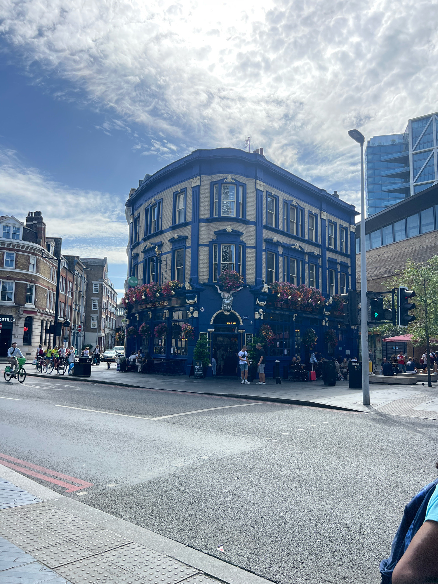 This is a wide, eye-level, outdoor, daytime photograph of a street corner in London, United Kingdom. The prominent subject is a traditional blue and cream-colored pub named "THE SHIPWRIGHT ARMS" on the corner of the intersection. The pub has ornate architectural details, including bay windows, decorative moldings, and hanging flower baskets overflowing with colorful blooms.
Several people are visible on the street and sidewalk. On the left side of the image, a man is riding a bicycle, and a group of people are gathered near a cluster of bicycles. In front of the pub, there are outdoor seating areas with patrons. Further down the street, there are more people and traffic, including cars and a bus. Modern glass buildings are visible in the background, contrasting with the historic architecture of the pub. A traffic light, a street lamp, and street signs are also present. The sky is partly cloudy with patches of blue. The street is paved and appears to be wet in some areas, suggesting recent rain. A pedestrian crossing with tactile paving is visible in the foreground.