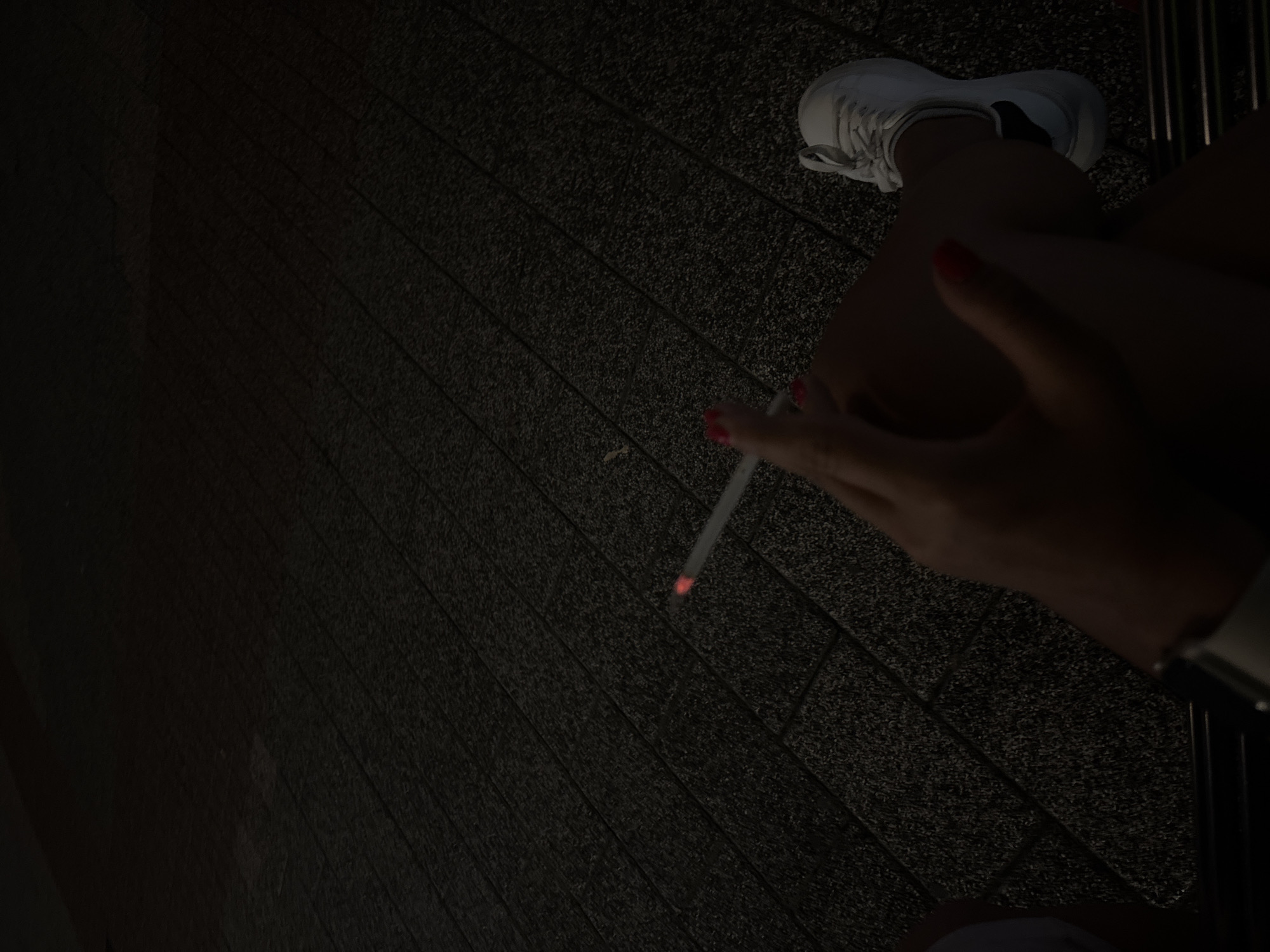 An individual is seated outdoors at night, holding a lit cigarette with their right hand. Their fingernails are painted red. They are wearing white sneakers. The ground is paved with dark rectangular tiles. The scene is dimly lit, suggesting an urban environment in Plovdiv, Bulgaria, possibly in a public seating area.