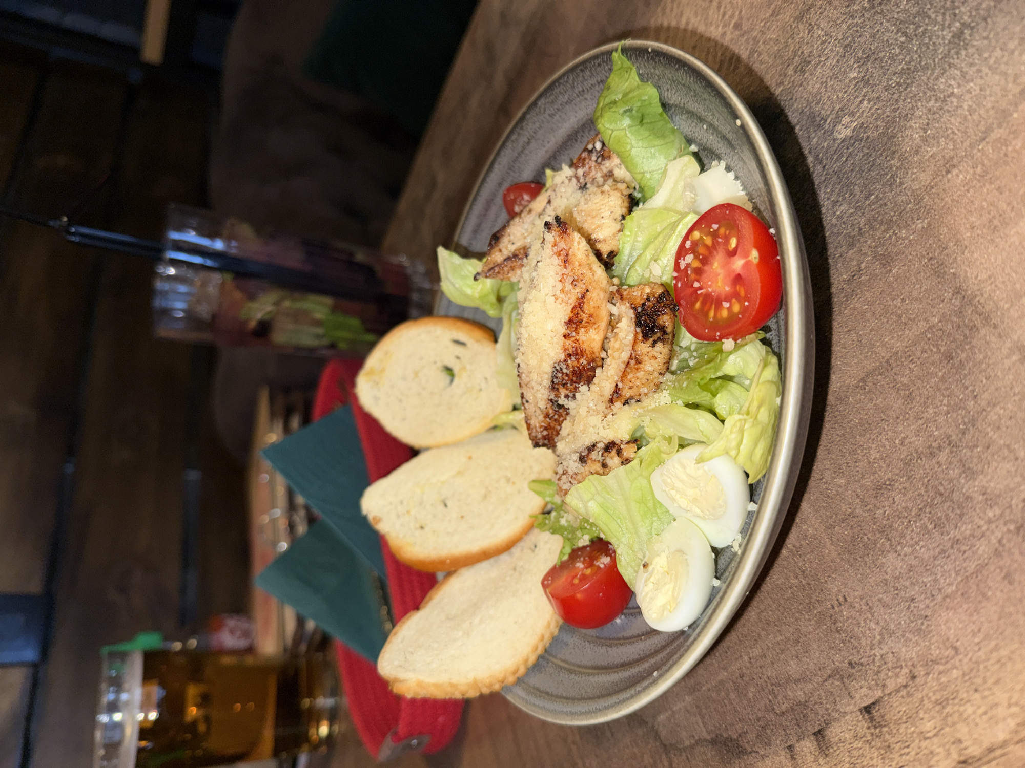 The image presents a close-up, slightly angled view of a Caesar salad served on a dark gray plate, positioned on a textured brown table. The main components of the salad include several pieces of grilled chicken breast, generously sprinkled with grated Parmesan cheese, fresh green and lighter-colored lettuce leaves, two halved cherry tomatoes, and two halves of hard-boiled quail eggs.
Alongside the salad, three elongated slices of toasted bread, likely baguette or similar croutons, are arranged on the plate. Underneath the plate, a dark blue-green folded napkin and a red fabric, possibly another napkin or placemat, are visible.
In the blurred background to the left, a tall glass containing a dark beverage with a black straw can be seen. The overall lighting is dim and ambient, suggesting an evening or nighttime setting within a restaurant or cafe environment. No people are visible in the frame. There is no visible text in the image. The scene captures a prepared meal, ready to be eaten, in a dining establishment, possibly in Chaikovsky, Russia, as per the provided context.