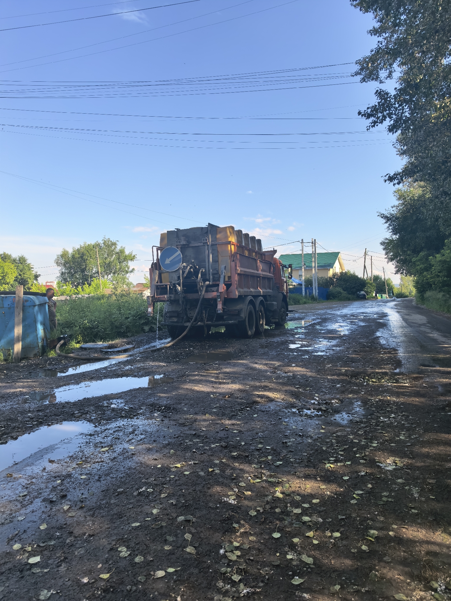 A truck with a large orange container is parked on a muddy, unpaved road. A hose is connected from the truck to a blue structure. Water appears to be flowing from the truck towards the blue structure, or possibly being pumped from it. A young boy in a camouflage outfit stands near the blue structure, looking towards the truck. The road is covered in puddles and fallen leaves. In the background, there are houses with green roofs, trees, and utility poles with power lines crisscrossing the sky. The sky is clear and blue, suggesting it is a sunny day. The scene appears to be in a rural or semi-rural area.