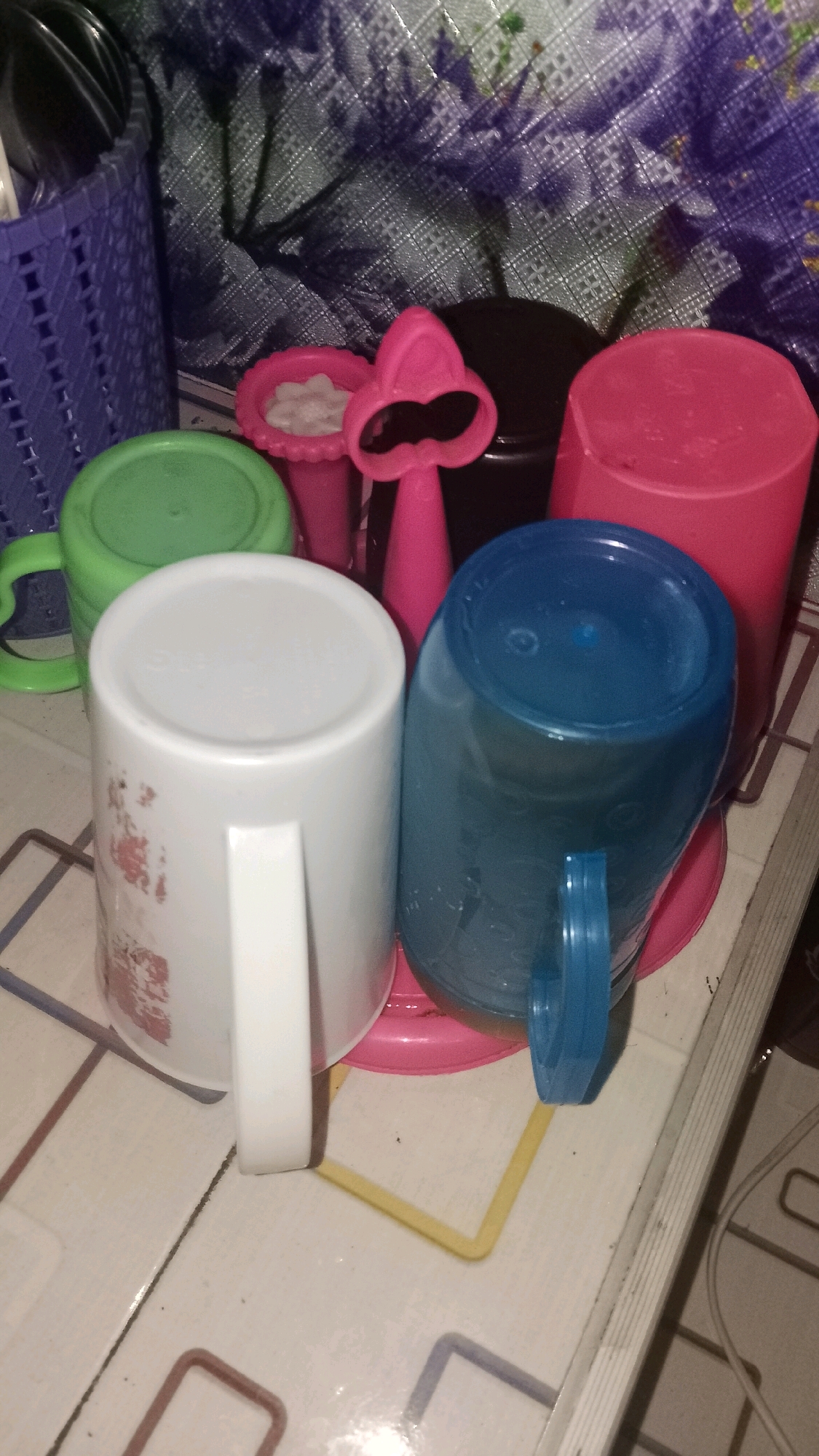 The image shows a close-up of several plastic mugs and some toys on a tiled surface. In the foreground, a white mug with a handle is positioned to the left, with a pattern of red floral designs visible on its side. To its right is a blue plastic mug, also with a handle, and a portion of its top is visible. Behind these, a green mug with a handle is partially visible on the left. In the center background, there are pink toy accessories and a black container. To the right of the black container is a tall, pink plastic mug. A woven purple basket filled with utensils is on the far left of the image, and a patterned cloth covers the background. The scene appears to be indoors, likely in a kitchen or dining area, with the patterned tiling suggesting it could be in Mulyo Haji, Indonesia.