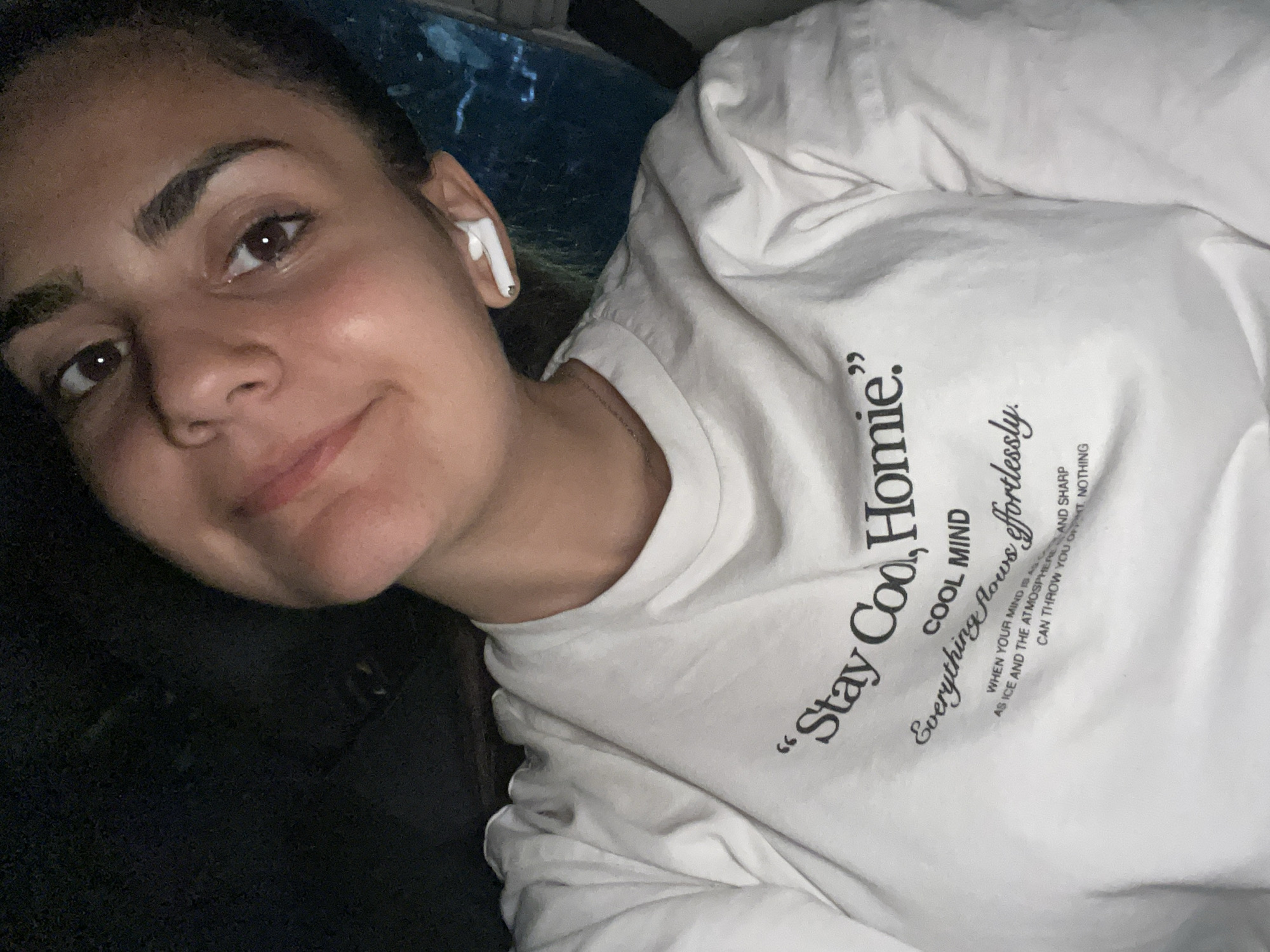 A young woman is posing for a selfie. She is wearing a white t-shirt with black text that reads "Stay Coo, Homie" and "Everything flows effortlessly". She has an AirPod in her left ear. Her expression is a slight smile. The background is dark and somewhat blurry. The location of the image is not specified.