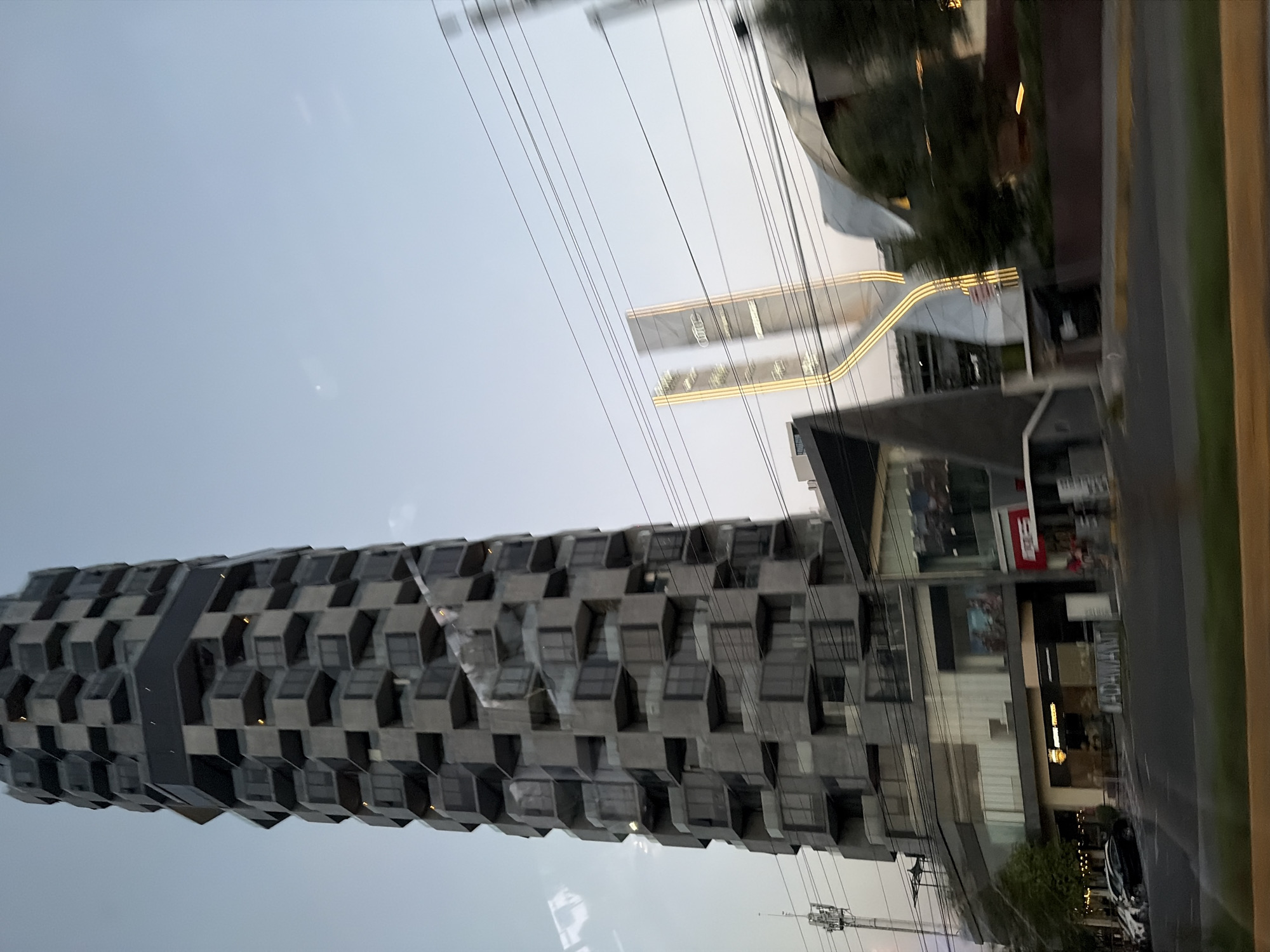 The image captures an urban streetscape, likely in San Bernardino Tlaxcalancingo, Mexico, under an overcast or hazy sky, suggesting either early morning, late afternoon, or a cloudy day.
The dominant subject is a tall, slender building on the left side, featuring a distinctive modern architectural style. Its facade is composed of numerous dark grey, geometric, and somewhat hexagonal or cube-like modules that protrude outwards, creating a textured, honeycomb-like appearance. These modules contain windows, some of which show internal illumination.
Multiple thin power lines crisscross the sky and mid-ground, partially obscuring other structures. Behind these lines, in the mid-ground, another modern building or complex is visible, characterized by a large, curved architectural element. This curve is accented by multiple horizontal lines of golden or warm yellow illumination. Further back, another section of a building also exhibits similar horizontal lighting.
In the foreground and to the right, a lower-rise commercial building features several storefronts with large glass windows. Faint text is visible on some of these facades, including "FORMAN" and possibly parts of words like "RED" or "ED" on a red sign. A vehicle, appearing to be a white or light-colored car, is parked on the street adjacent to this commercial building. A section of a road or pavement runs along the far right edge of the frame. Some green foliage and trees are scattered throughout the scene, with reflections visible on building surfaces. The scene is static, showing no specific human activity.