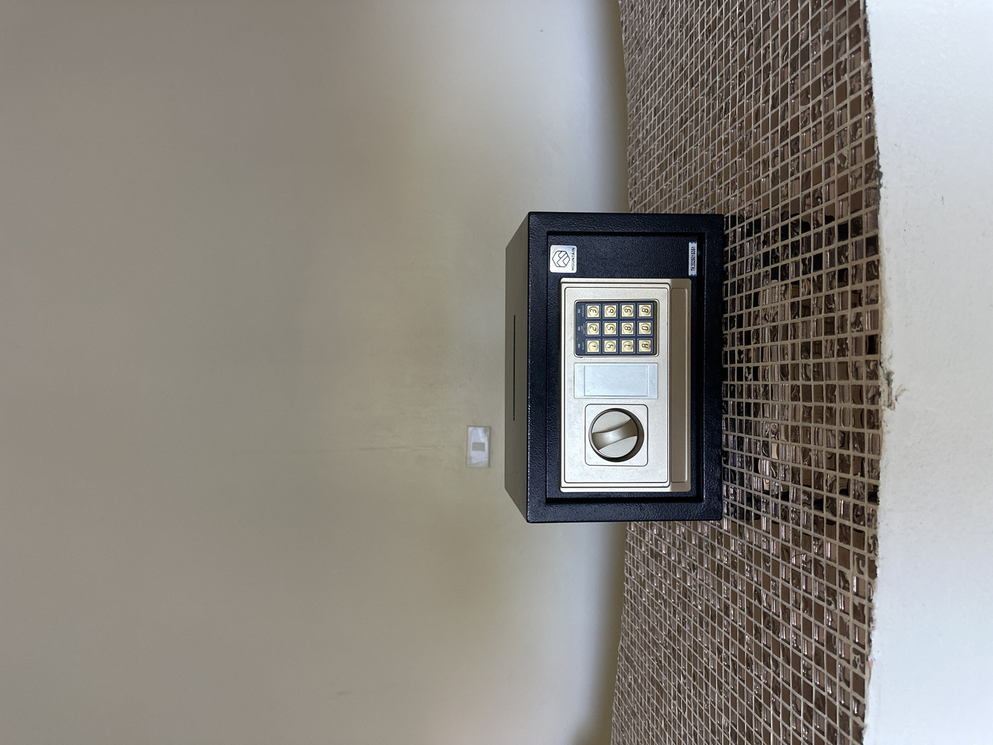 A black safe with a digital keypad and a rotary dial sits centered on a mosaic-tiled surface in front of a plain, beige wall. The safe has a slot on its top surface. A small logo is visible on the right side of the safe's door, and a serial number is printed below it. The wall behind the safe has a light switch on it. The scene appears to be indoors, likely in a room or a similar enclosed space. The location is specified as Koh Phangan, Thailand.