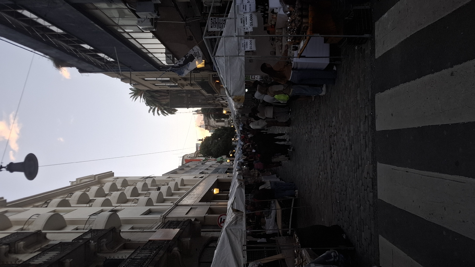 The image depicts a bustling street market in Buenos Aires, Argentina. The street is lined with stalls, each displaying handcrafted items and local products. People are seen browsing and interacting with vendors, suggesting a vibrant commercial atmosphere. Buildings with intricate architectural details frame the street, reflecting the city's historical character. A crosswalk runs across the street, adding to the urban landscape. The photo captures the cultural and economic activity that defines this area of Buenos Aires.