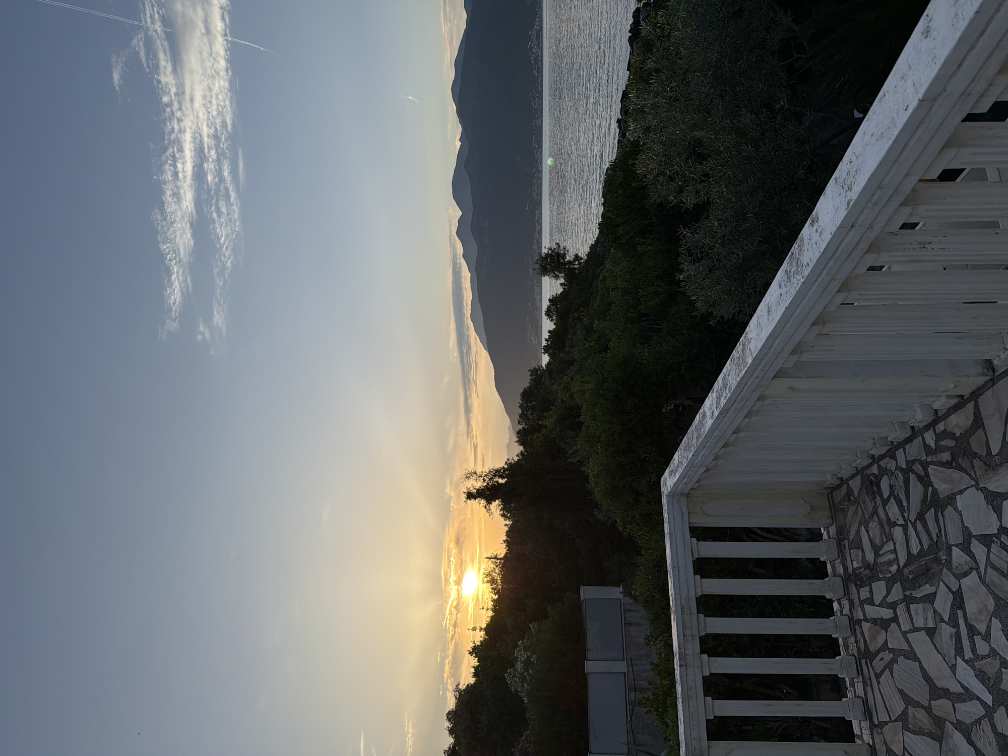 This image captures a serene sunset over a body of water, viewed from an elevated perspective, likely a balcony. The sky, transitioning from a clear blue at the top to warm orange and yellow hues around the setting sun on the left, features a long, wispy cloud and a faint contrail in the upper left.
Silhouetted against the glowing horizon are dark, undulating mountains that extend across the middle of the frame, separating the sky from the expansive body of water. The water surface, likely the Adriatic Sea given the Gošići, Montenegro location, shimmers with reflected light, appearing silvery-grey on the right side of the frame.
In the mid-ground, dense, dark green trees and bushes are silhouetted against the sunset and water, partially obscuring the sun's brilliance. Below this foliage, some lighter-colored, rectangular structures, possibly air conditioning units or water tanks, are visible, suggesting a building below the viewpoint.
The foreground features a white balcony railing with vertical slats, occupying the lower right portion of the frame. The balcony floor, visible to the right of the railing, is paved with irregular, light-colored stone tiles, and an adjacent wall covered in similar tiles is partially visible on the far right. The time of day is clearly sunset, creating a peaceful and tranquil atmosphere. No people or text are visible in the image.