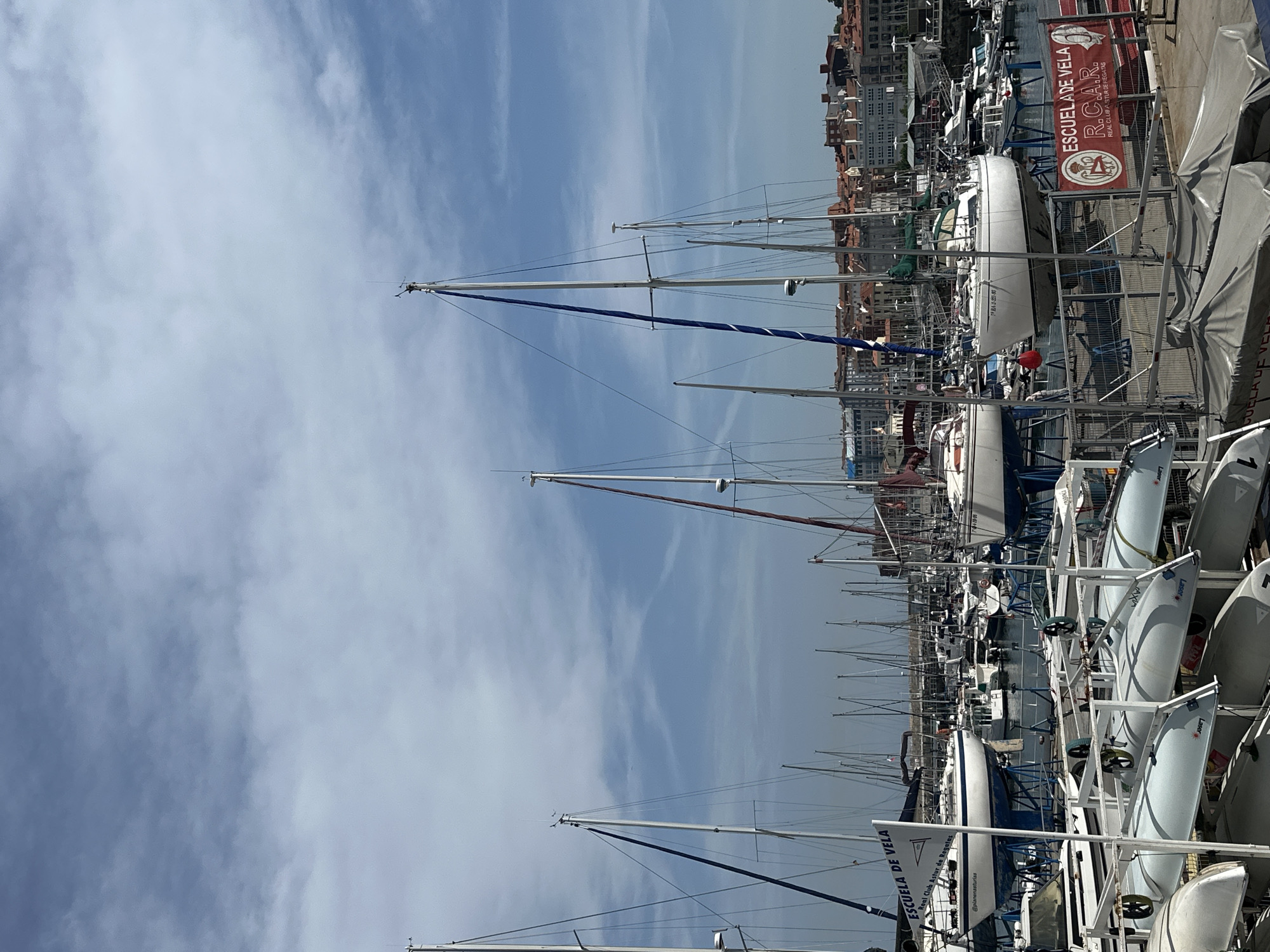 The image depicts a marina in Gijón/Xixón, Spain. A multitude of sailboats with tall masts dominate the frame, clustered tightly together in the harbor. Smaller dinghies are neatly stacked on a metal rack, displaying the word "Laser". A sign reads "ESCUELA DE VELA Real Club Astur de Regatas". Buildings are visible in the background. The sky above is partly cloudy.