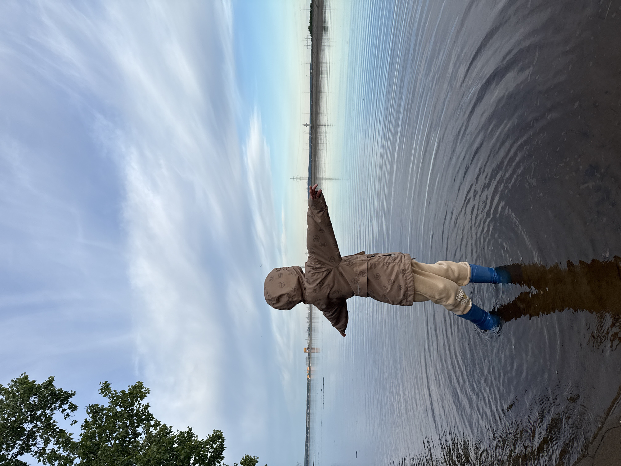 A child stands in a body of water near Noviy, Russia. The child wears a brown coat, light-colored pants, and blue rubber boots. Ripples emanate from the child's legs in the water. The sky is partly cloudy. Power lines can be seen across the water in the distance. Trees are visible on the left.