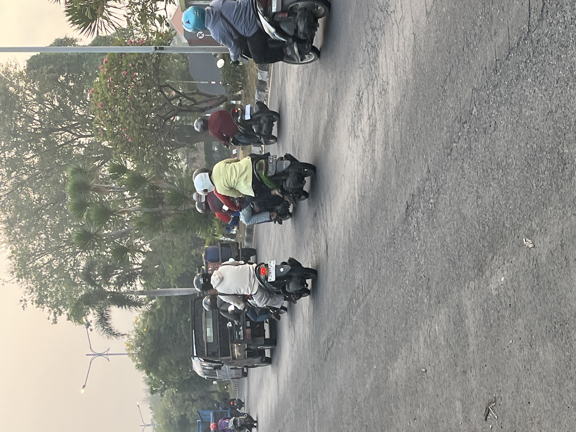 A group of motorcyclists and a utility truck are on a paved road in Kludan, Indonesia, during daylight hours, possibly in the early morning or late afternoon given the soft light and hazy sky. The road has a cracked surface and is lined with trees, including palm trees, and vegetation. In the background, a street lamp with an ornate design is visible.
On the road, several people are riding motorcycles. One motorcyclist in the foreground, wearing a light-colored hoodie and grey pants, is riding a black scooter. Behind them, another person on a motorcycle wears a white helmet and a yellow shirt. Further back, a utility truck with an open bed is moving. Other motorcyclists are visible throughout the scene, some wearing helmets of various colors, including white, black, and blue. The environment appears to be a suburban or semi-rural area.